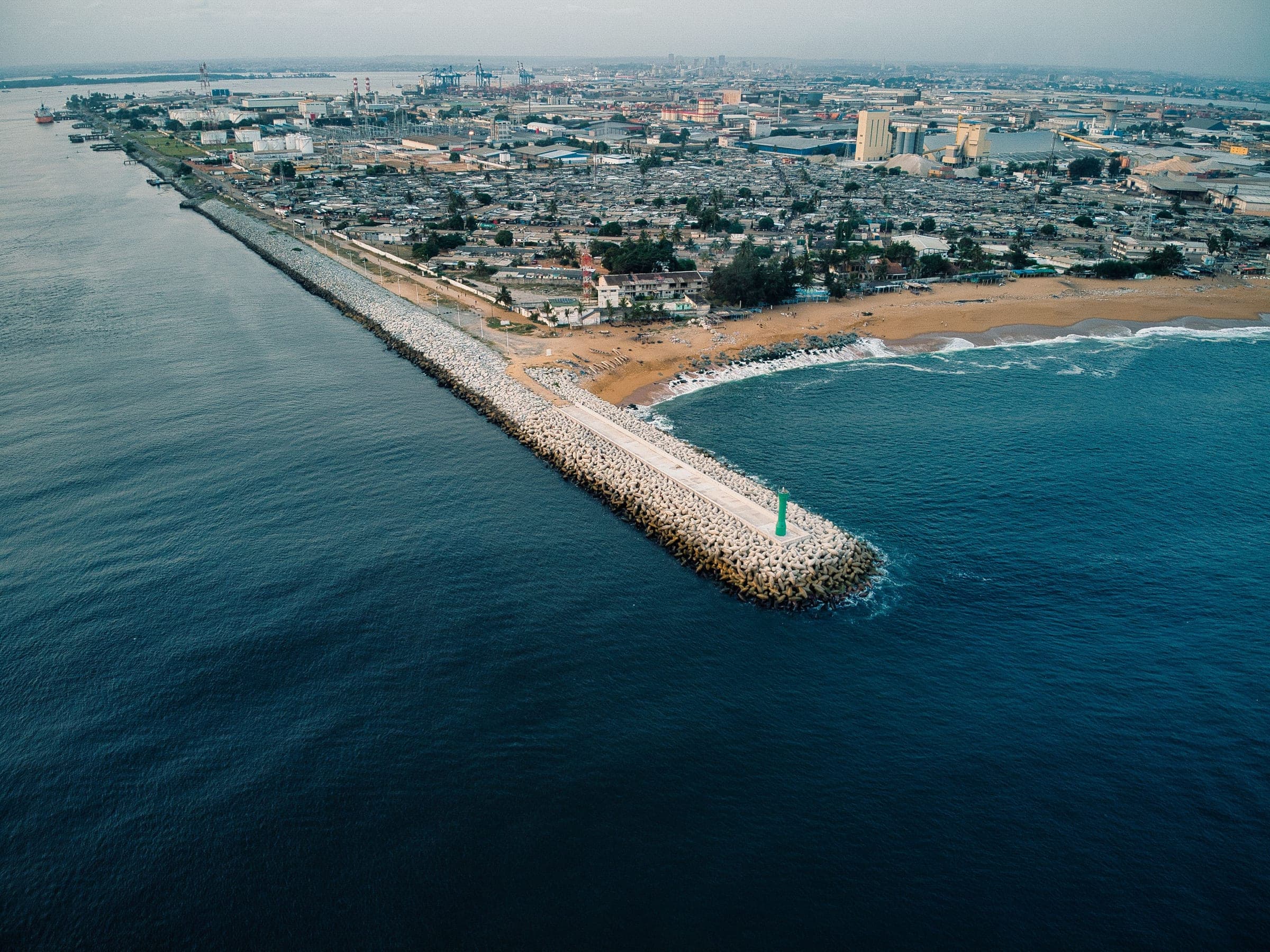 Aerial view of a long stone breakwater running out into dark blue water beside the port district of Abidjan, Côte d’Ivoire, with a sandy beach and low buildings behind it.