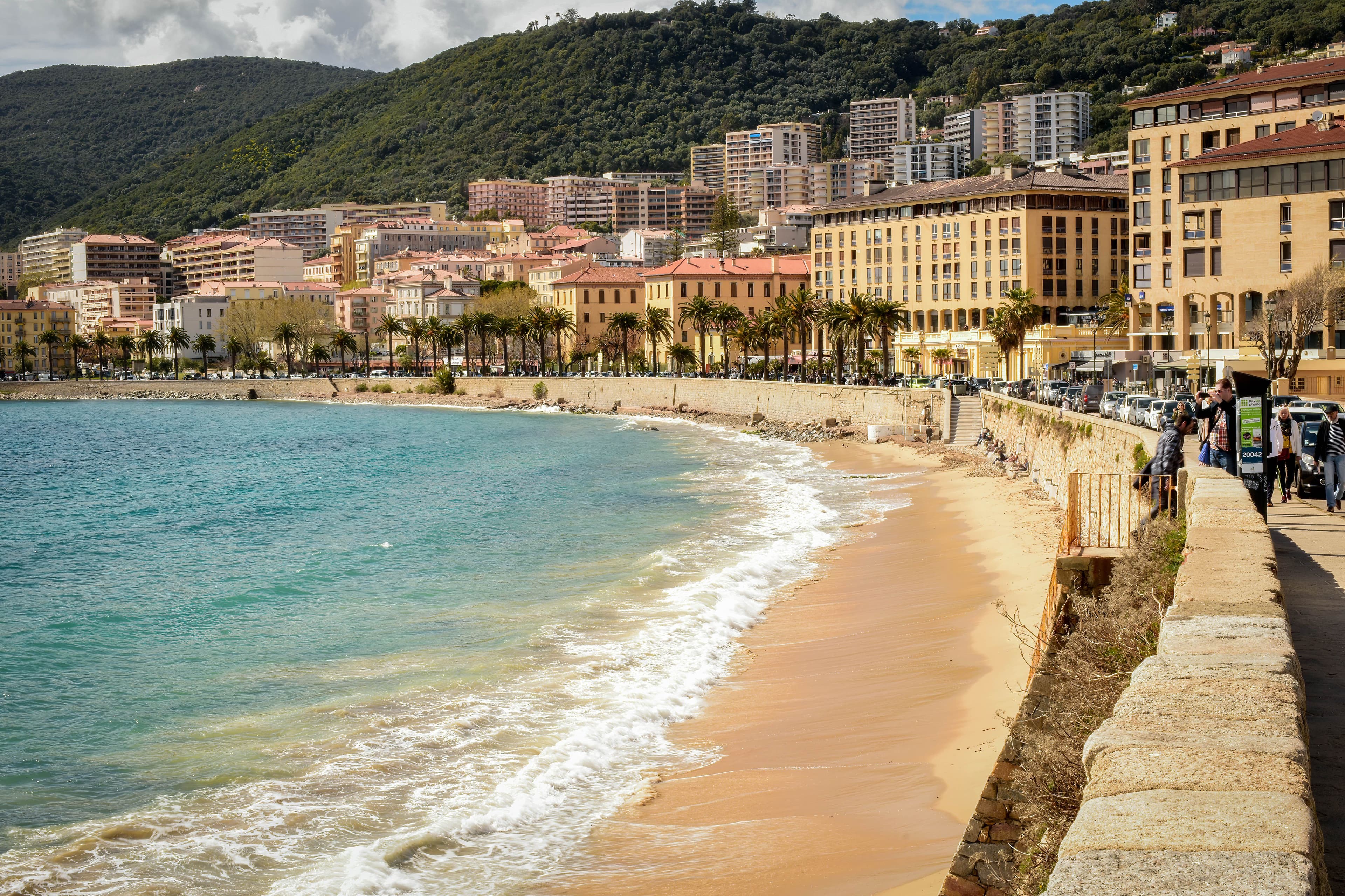 Ajaccio's town beach and sea wall below a palm-lined seafront boulevard, with apartment buildings and wooded hills behind. The marathon starts and finishes on the city's western seafront and runs out along this coast to La Parata.