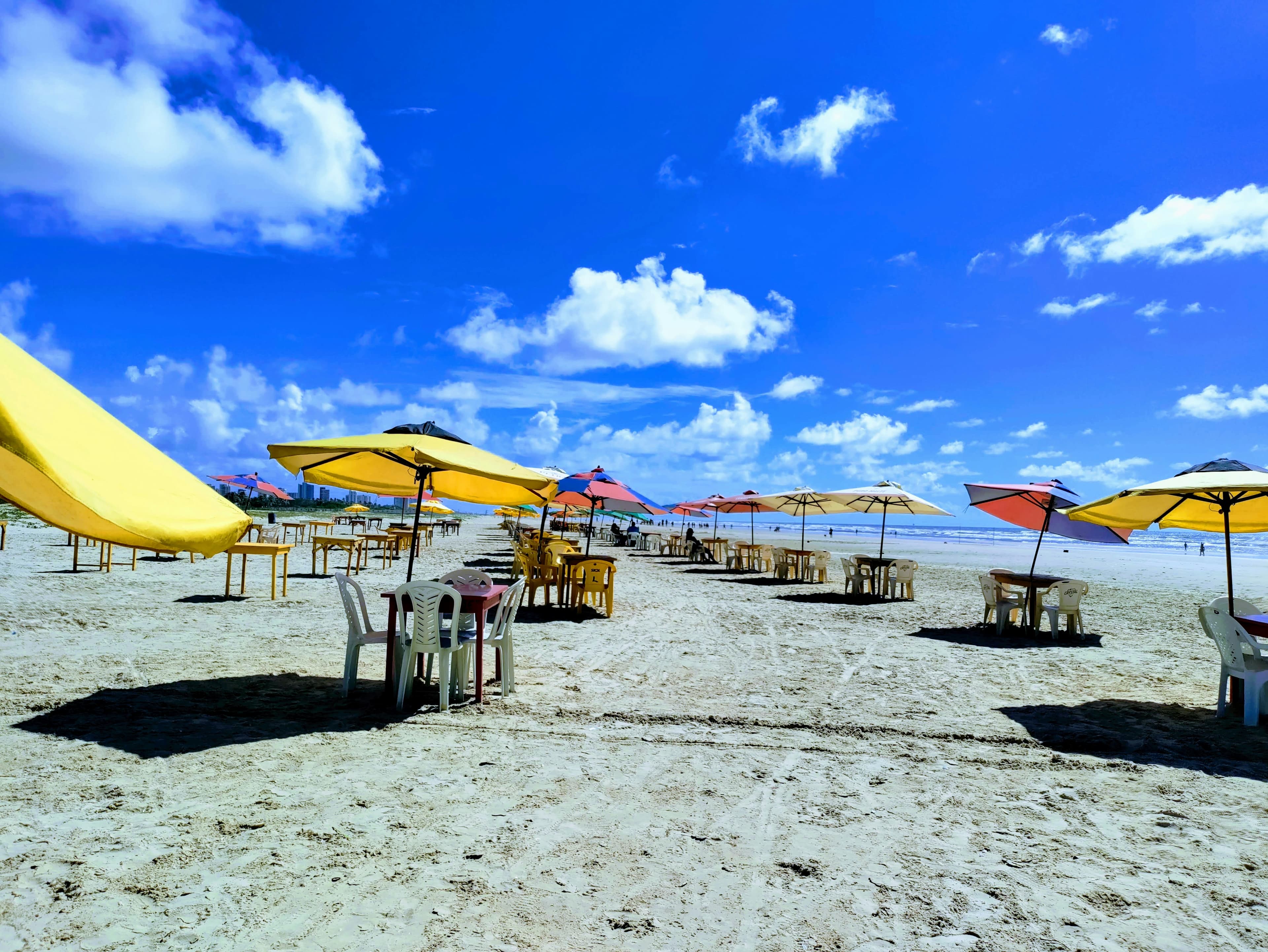 Rows of yellow and striped beach umbrellas with tables and chairs on a wide sandy beach under a deep blue sky, with the city skyline at the far end