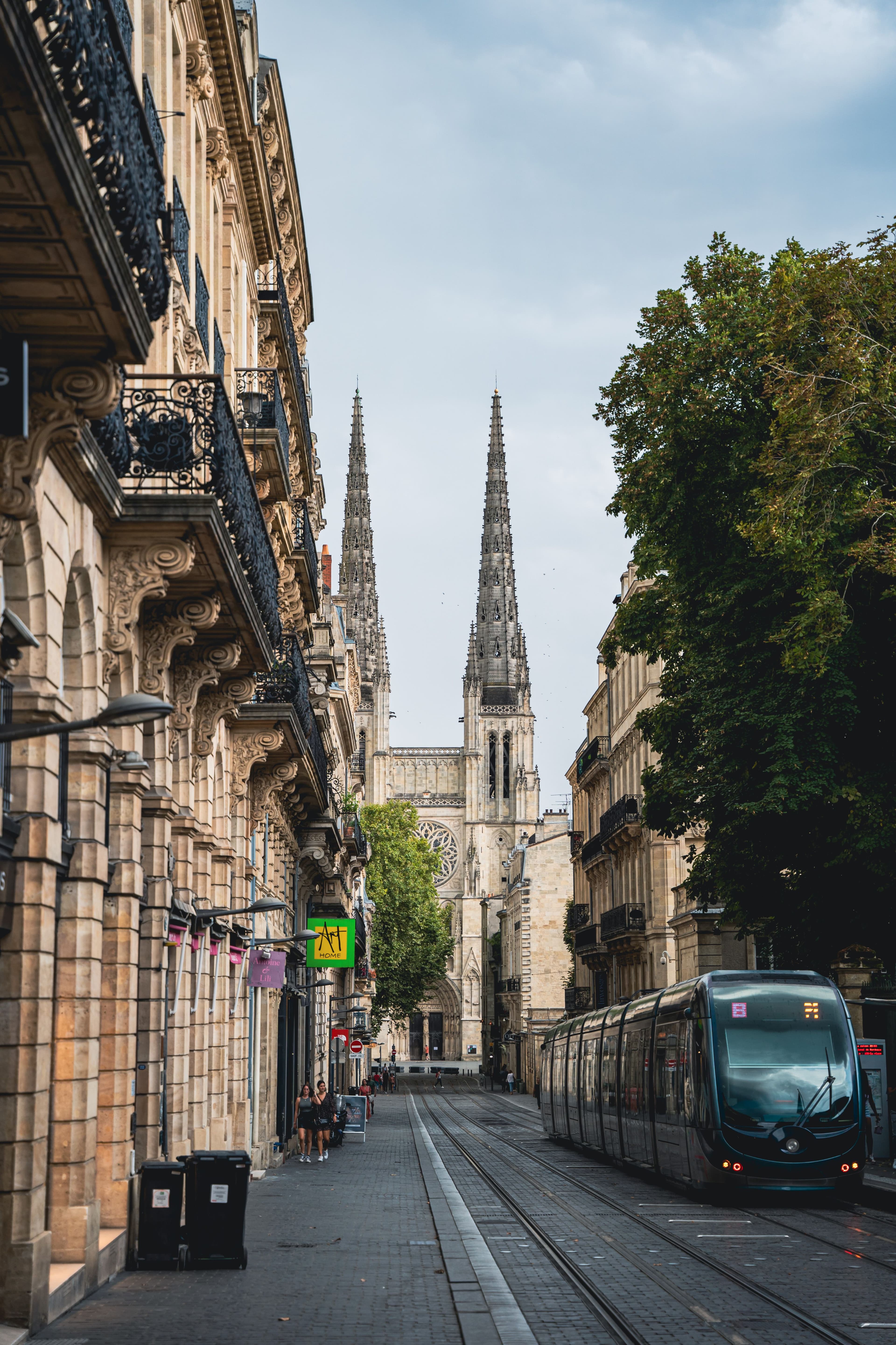 A TBM tram on the tracks in a Bordeaux street lined with 18th-century limestone facades and wrought-iron balconies, with the twin Gothic spires of the cathedral rising at the end of the street.
