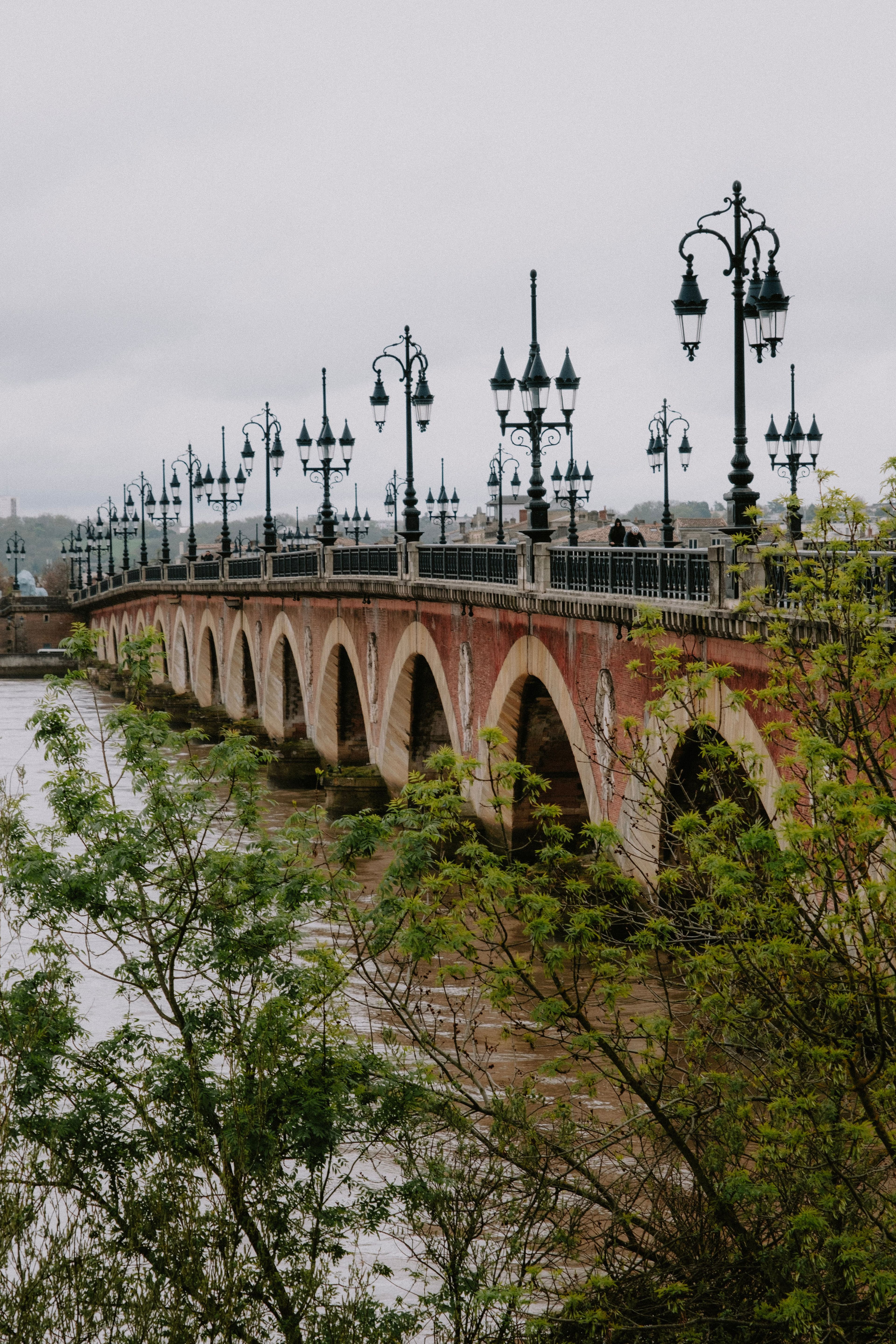 The Pont de Pierre in Bordeaux, a long brick-and-stone arched bridge carrying ornate cast-iron lamp standards across the wide, silt-brown Garonne under a grey sky.