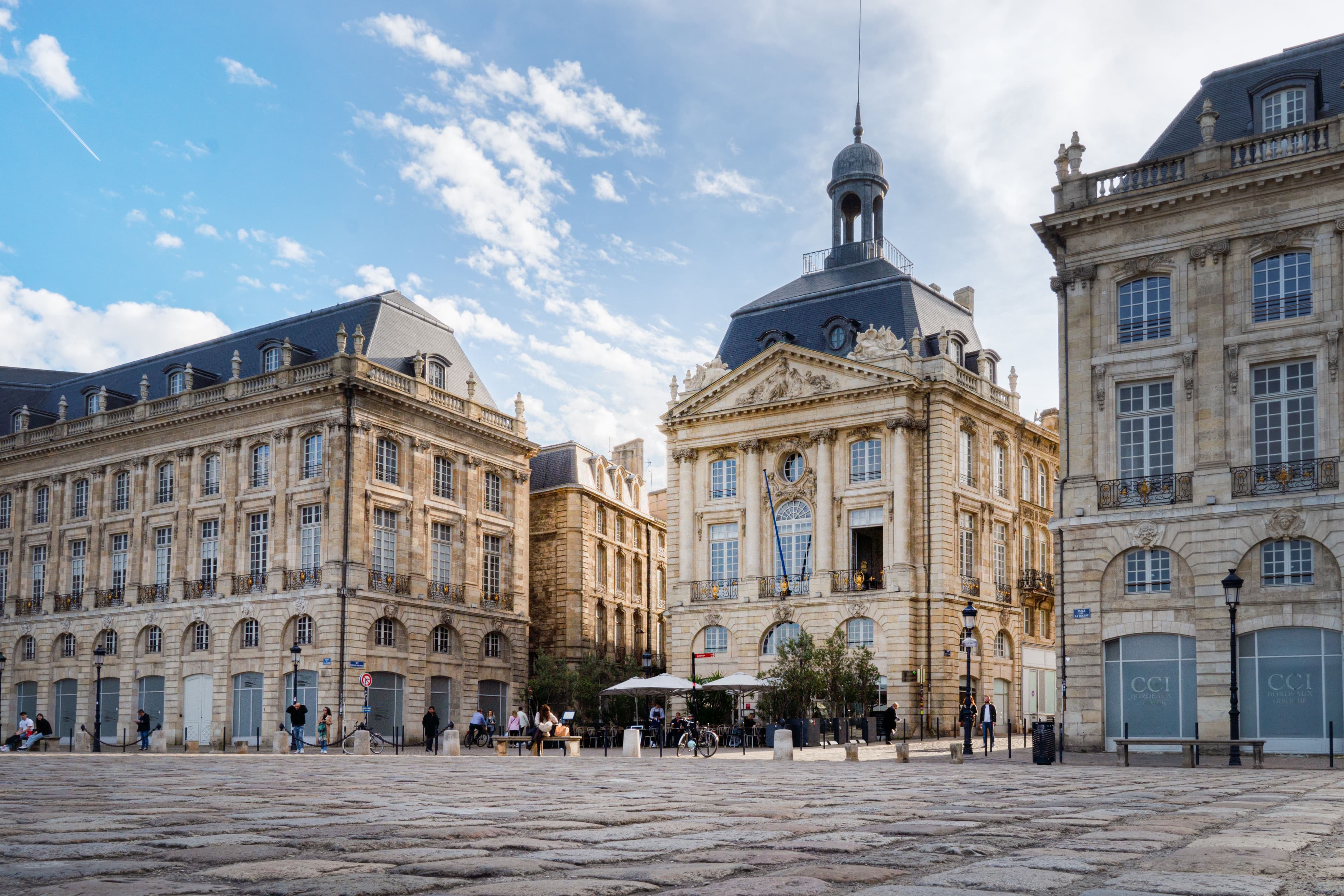 Place de la Bourse in Bordeaux, the 18th-century limestone square with its domed pavilion and the CCI building, seen across the open cobbles under a blue sky. The square is named among the marathon's course landmarks.
