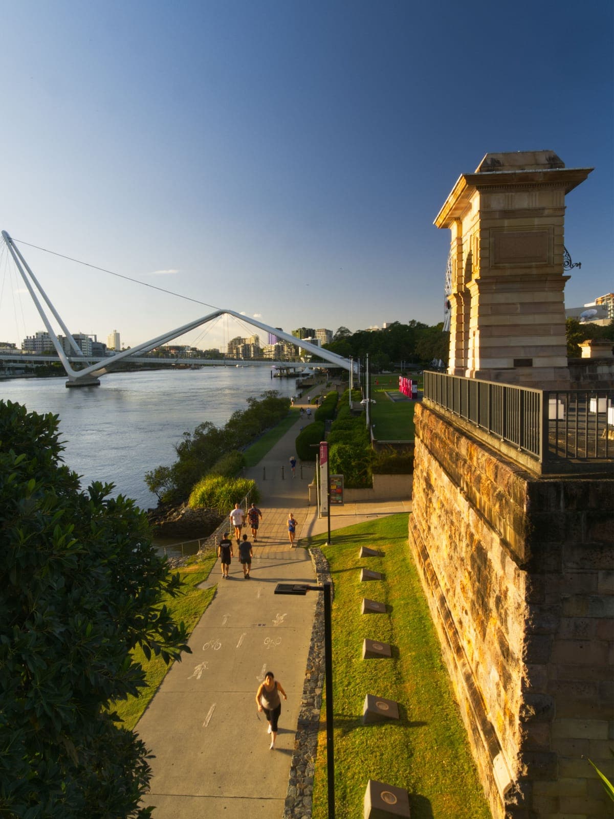 Runners on the riverside path below a sandstone pier on the Brisbane River at sunset, with the Kurilpa pedestrian bridge in the background