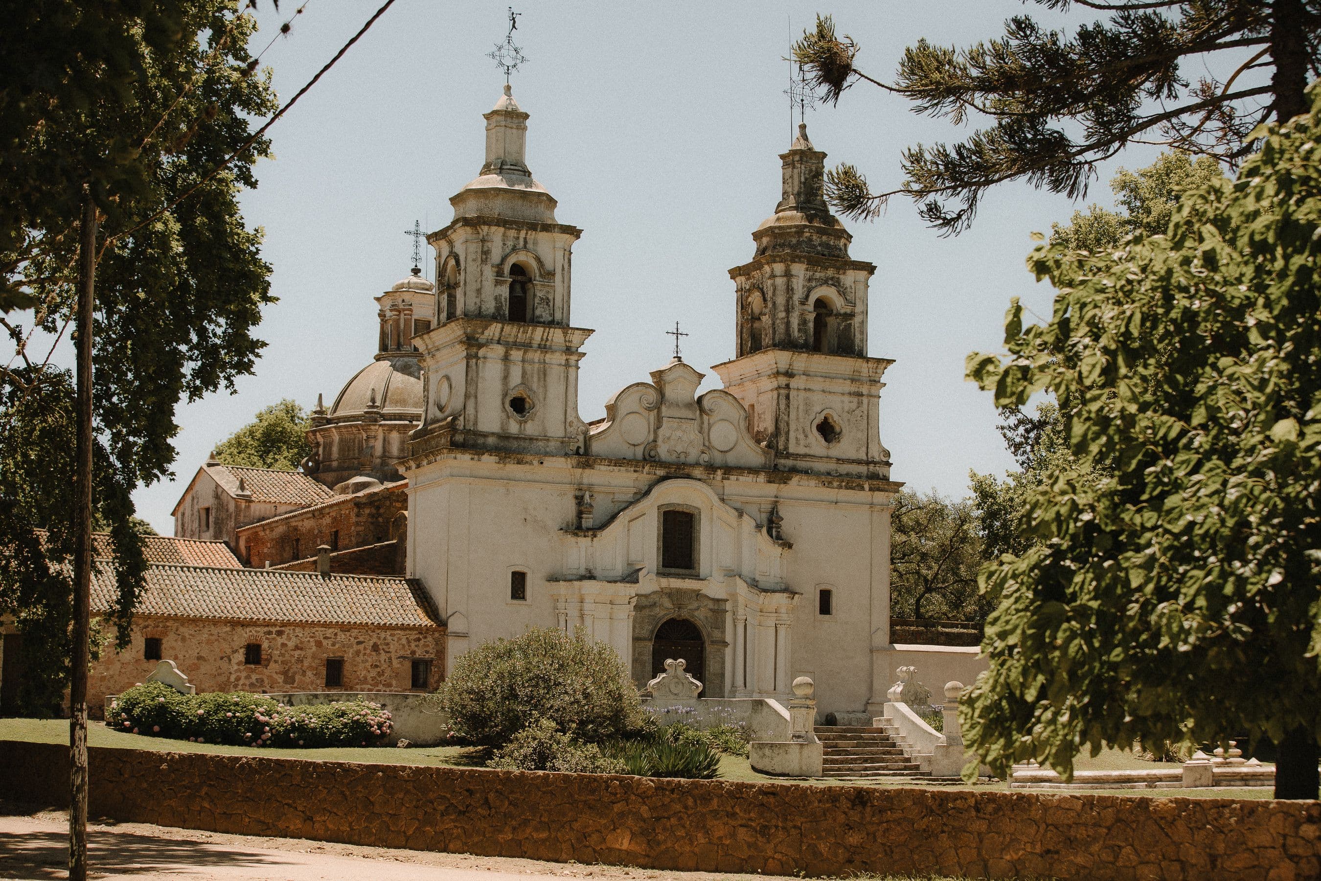 A whitewashed Jesuit estancia church with a bell tower and curved gable beside a stone wall and green lawn under a blue sky near Córdoba, Argentina