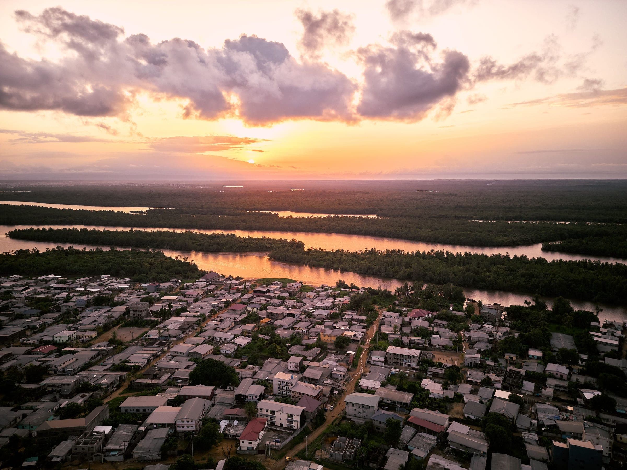 Aerial view at sunset of a Douala neighbourhood of low roofs beside a wide river, with dark mangrove forest on the far bank and a pink and orange sky over scattered clouds.