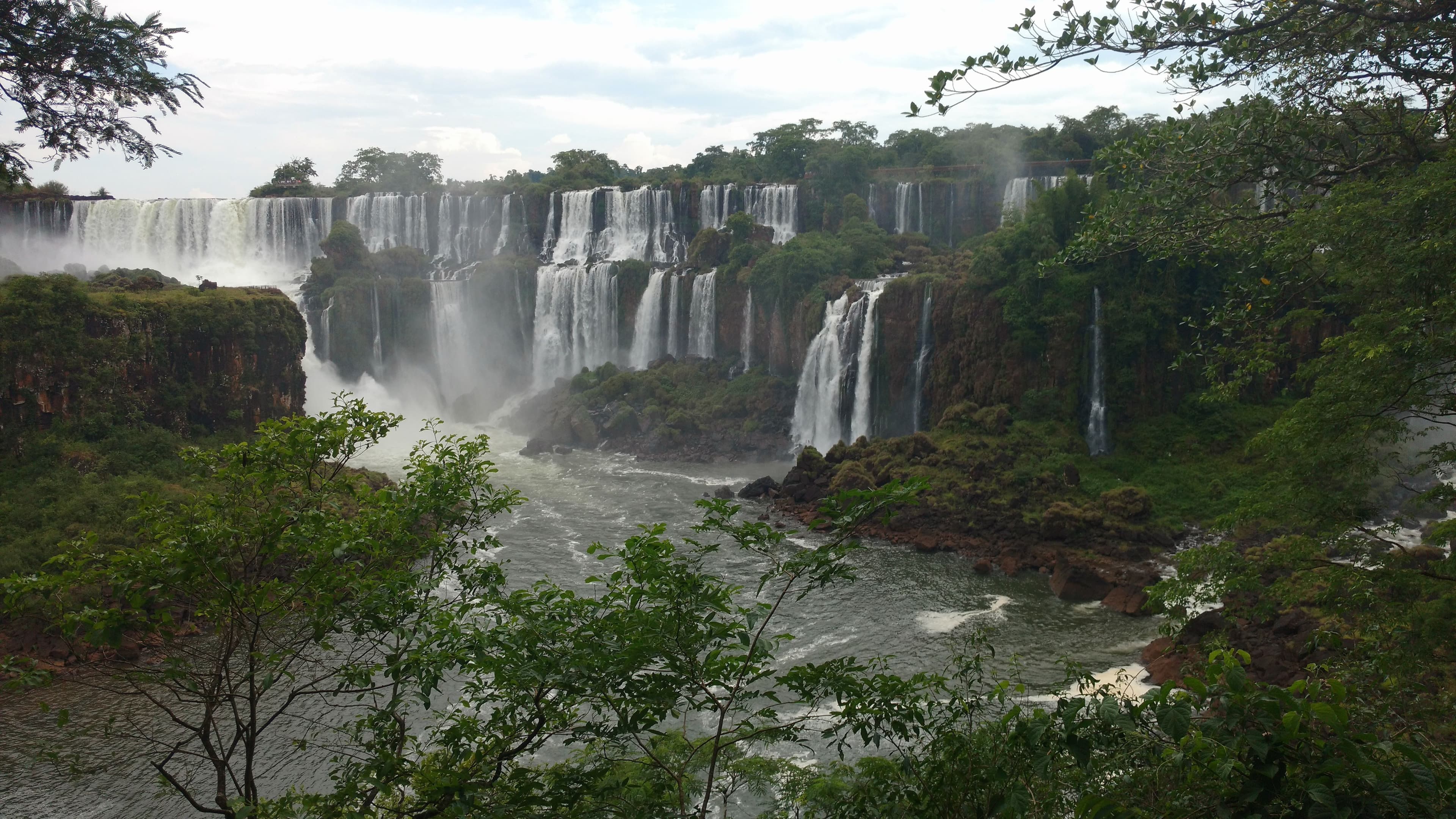 Iguazu Falls: curtains of white water pouring over a wide basalt cliff edge into the forested gorge beneath a blue sky