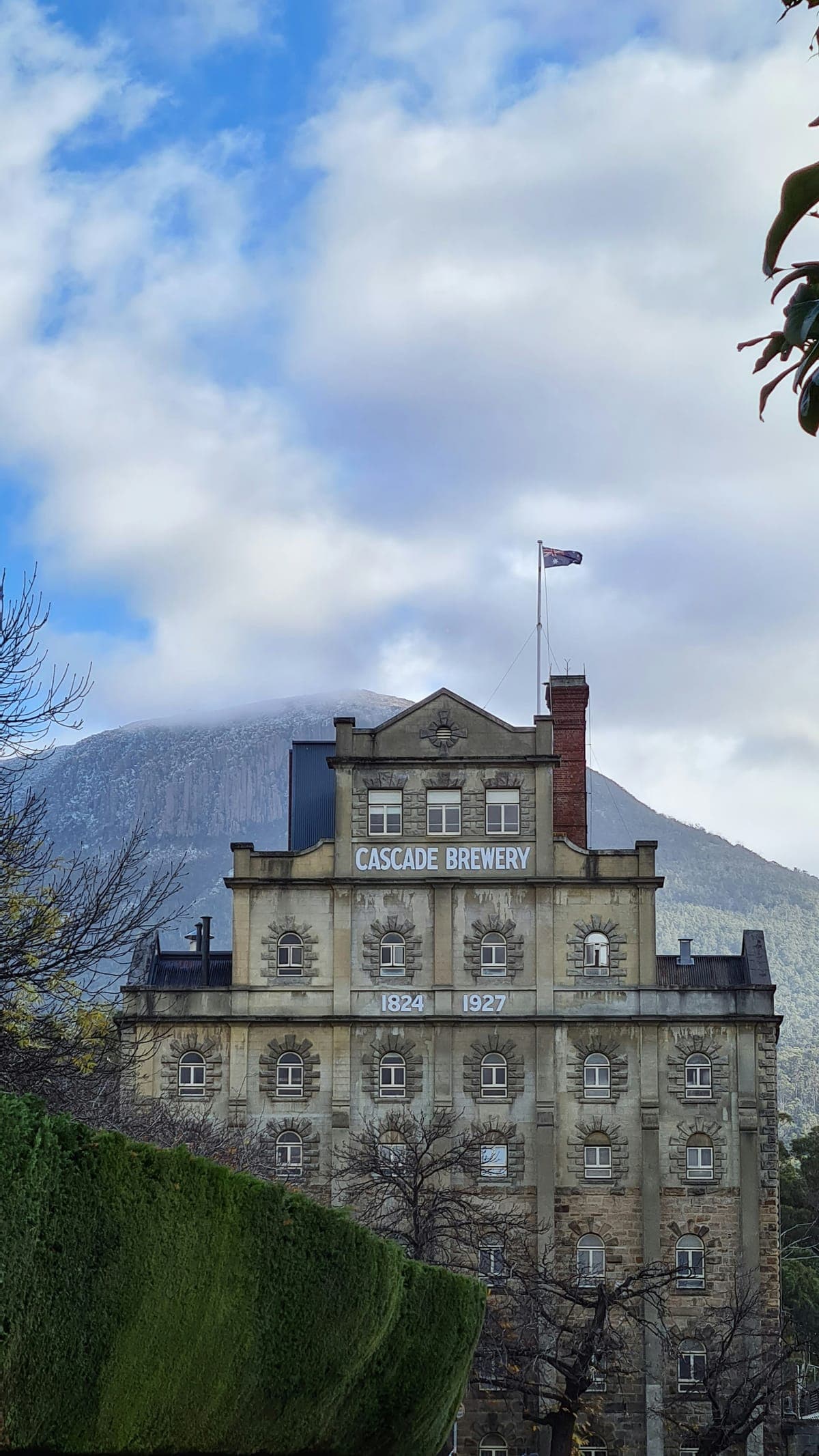Cascade Brewery in South Hobart, dated 1824 and 1927 on its facade, with kunanyi / Mount Wellington behind it under cloud