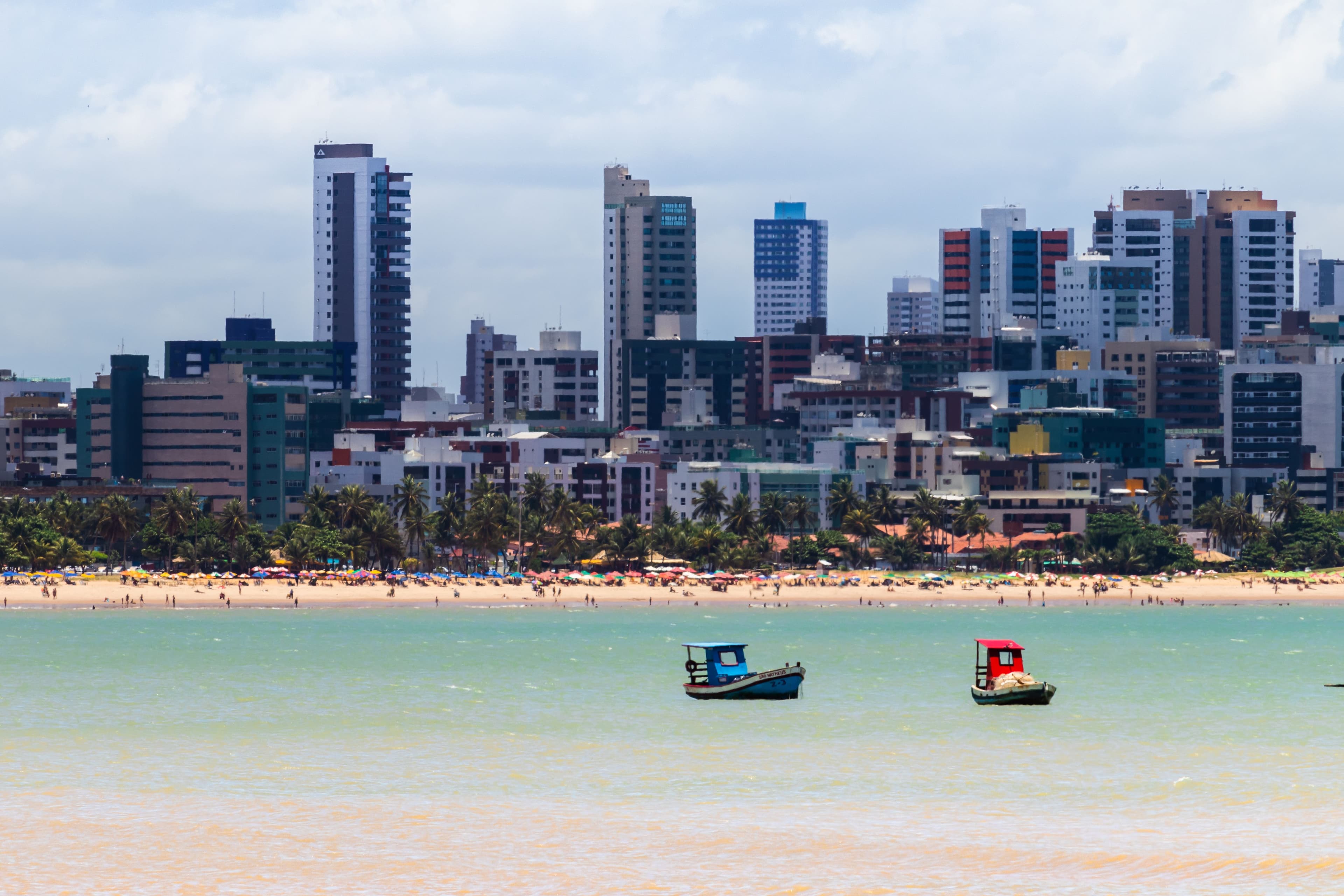 Moored boats in the foreground with the João Pessoa skyline and waterfront beyond, under a pale sky