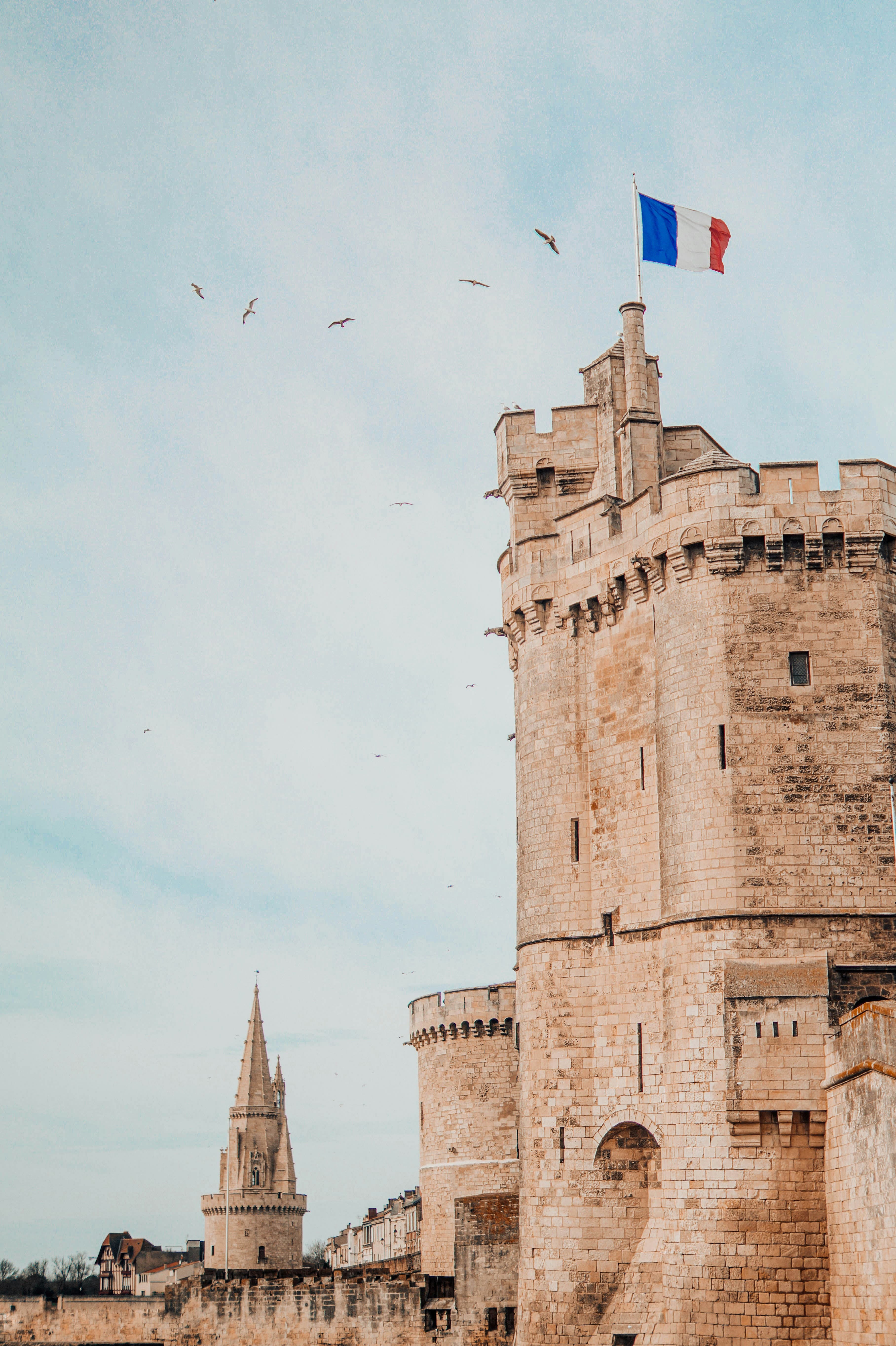 The Tour Saint-Nicolas at La Rochelle, a massive crenellated medieval harbour tower flying the French flag, with the spire of the Tour de la Lanterne behind it and gulls overhead. The marathon finishes beside these towers.