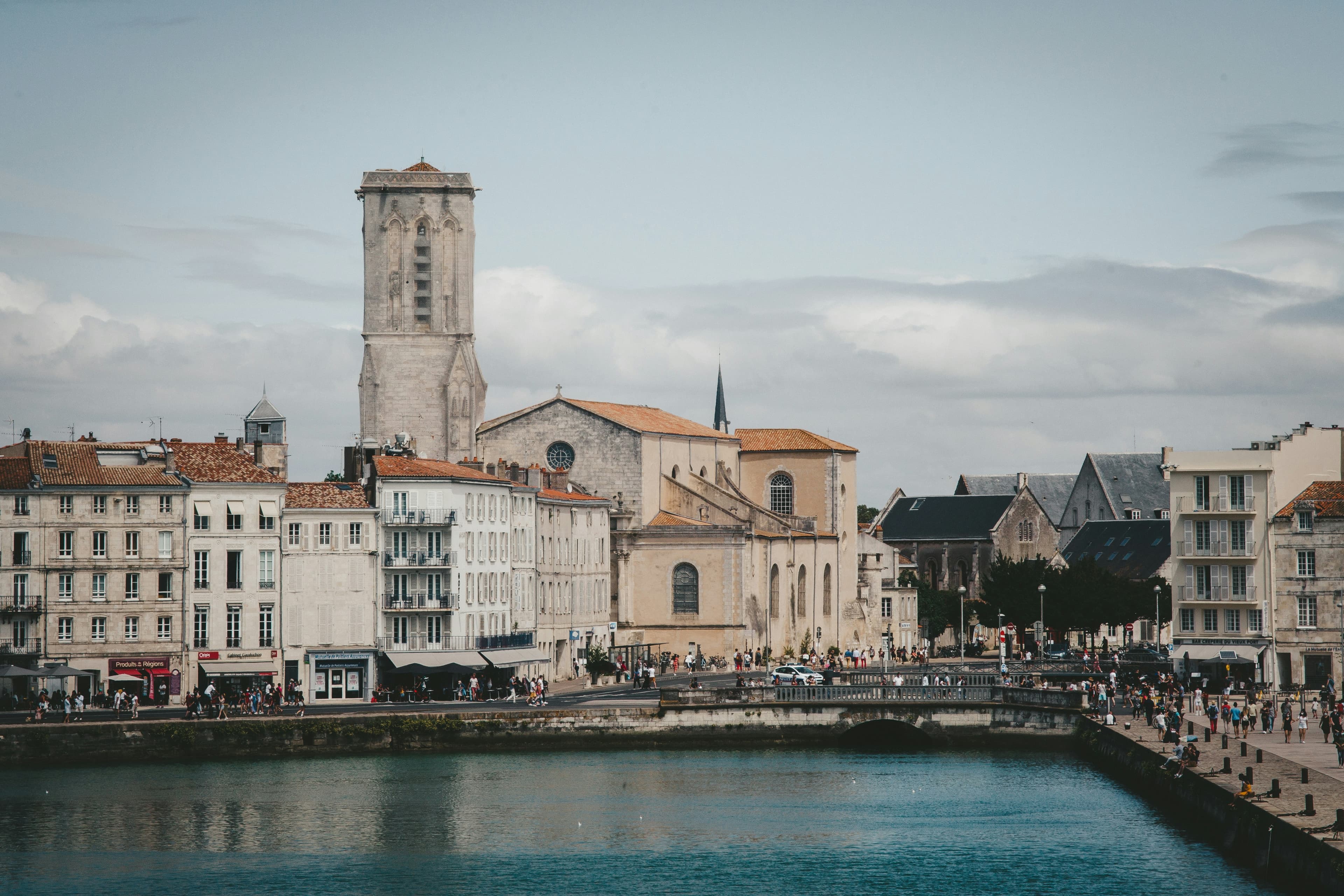 The Vieux-Port quayside at La Rochelle, with pale stone townhouses, cafés and the tower of the Église Saint-Sauveur rising behind, seen across the harbour water. The marathon finishes beside this harbour.