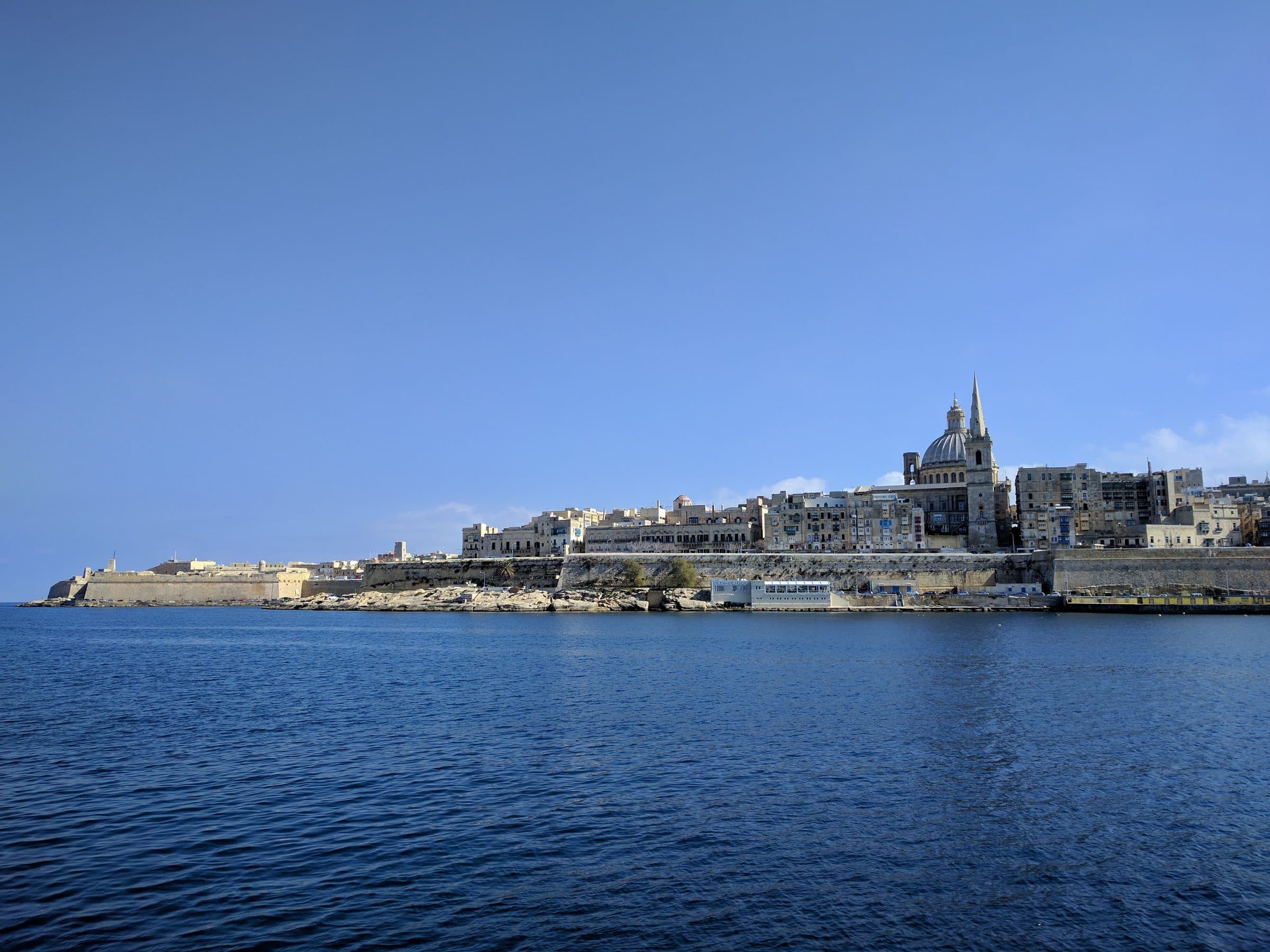The Valletta skyline across the harbour on a clear day, with the dome and spire of the Carmelite church above honey-coloured bastions and Fort St Elmo at the end of the peninsula on the left, over deep blue water