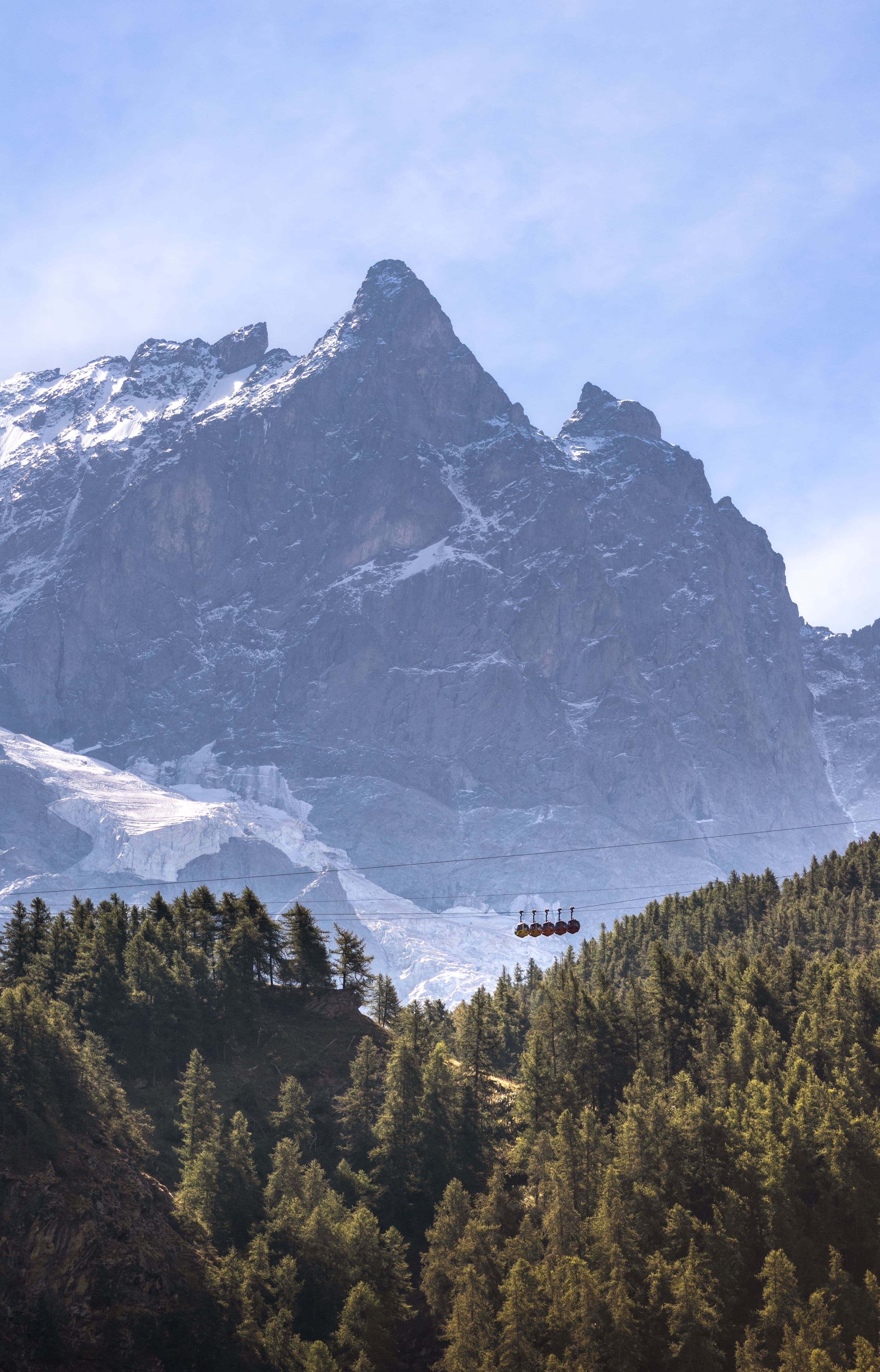 A sharp rock peak above a glacier tongue in the Chamonix valley, with a small red cable car crossing above a forested ridge in the foreground, showing the scale of the terrain the 42km course crosses