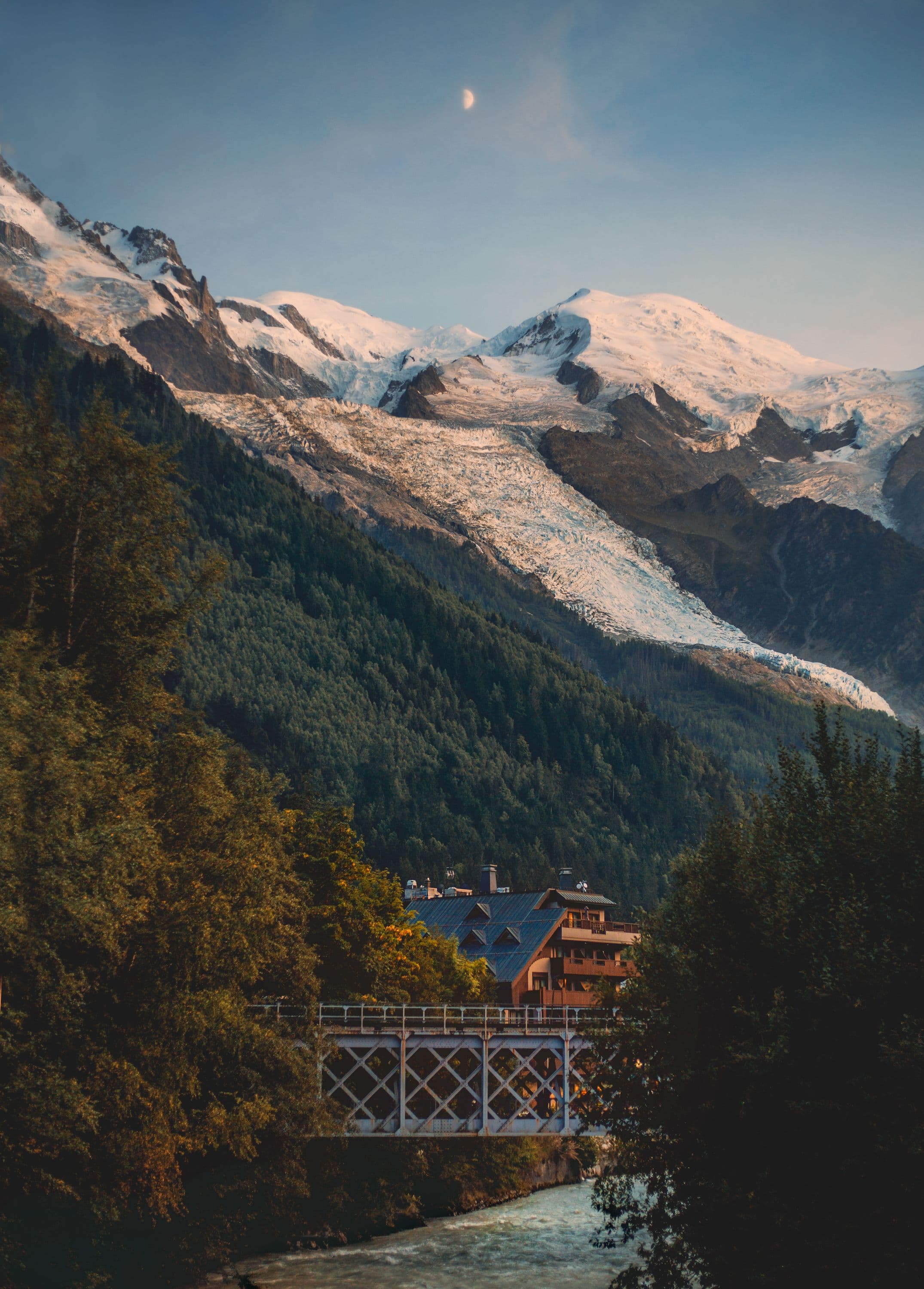 A glacier spilling down towards the wooded Chamonix valley at dusk with a crescent moon above, a chalet and a lattice iron railway bridge over the Arve in the foreground, close to the race start and finish in the town