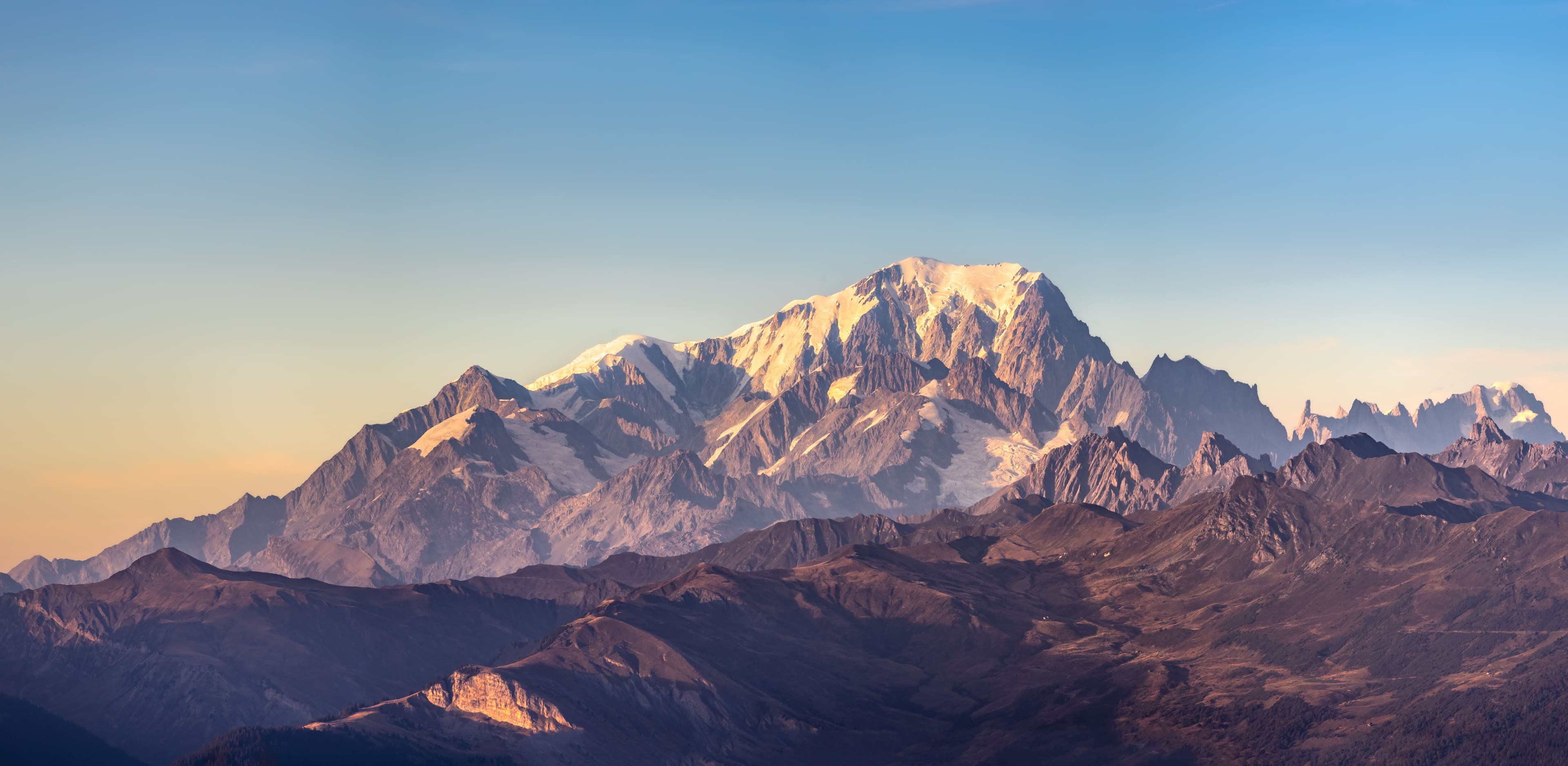 The snow-covered summit ridge of the Mont Blanc massif catching low sun above a long line of darker foreground ridges, the range the 42km du Mont-Blanc runs beneath from Chamonix