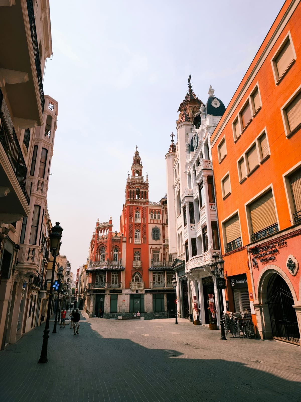 A pedestrian street in the Badajoz old town lined with orange and white facades, ending at a red neo-Mudéjar tower building with a tiered belfry, in the historic centre the marathon climbs through after km 30