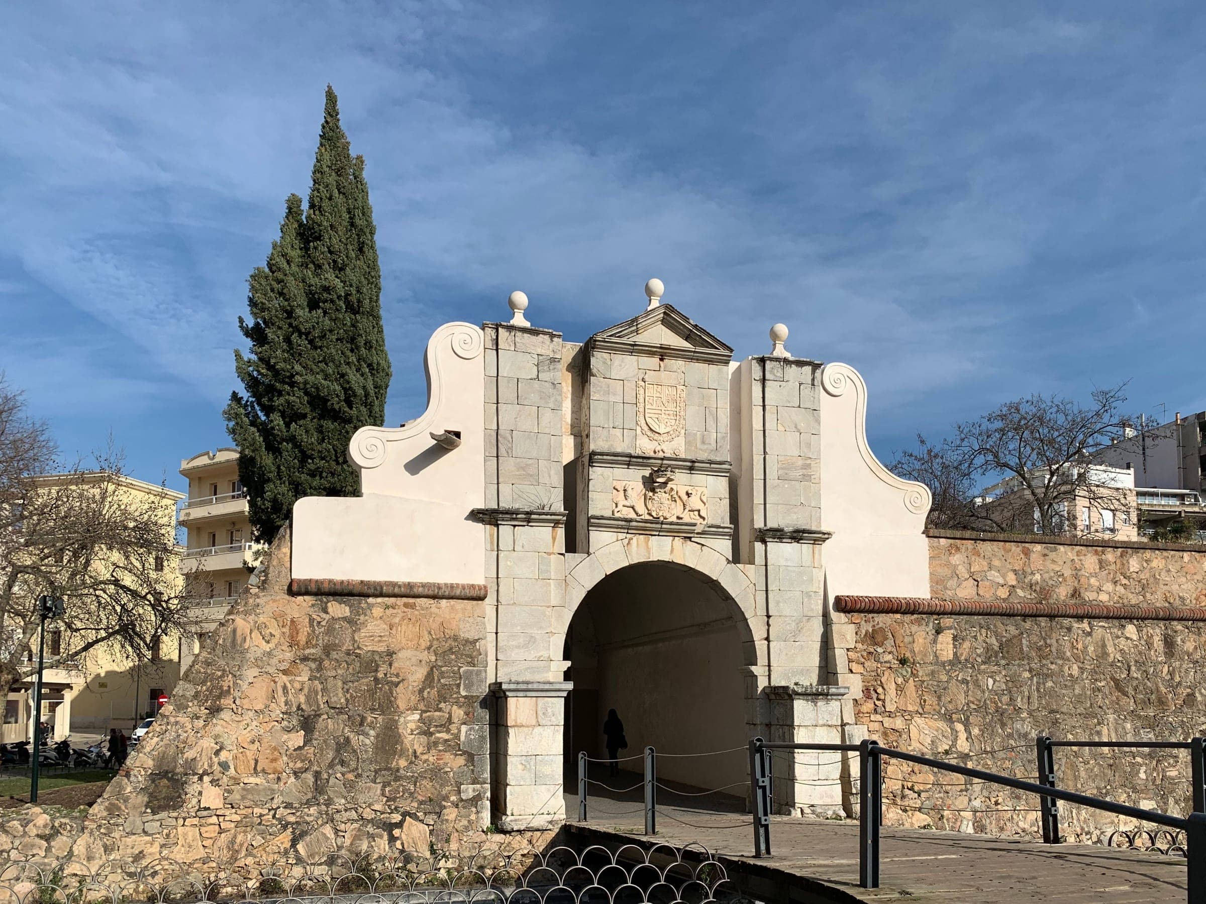 A gateway in the city walls of Badajoz: a stone arch under a pediment carved with the royal coat of arms, flanked by white scrolled gables, with a footbridge crossing to it past a tall cypress