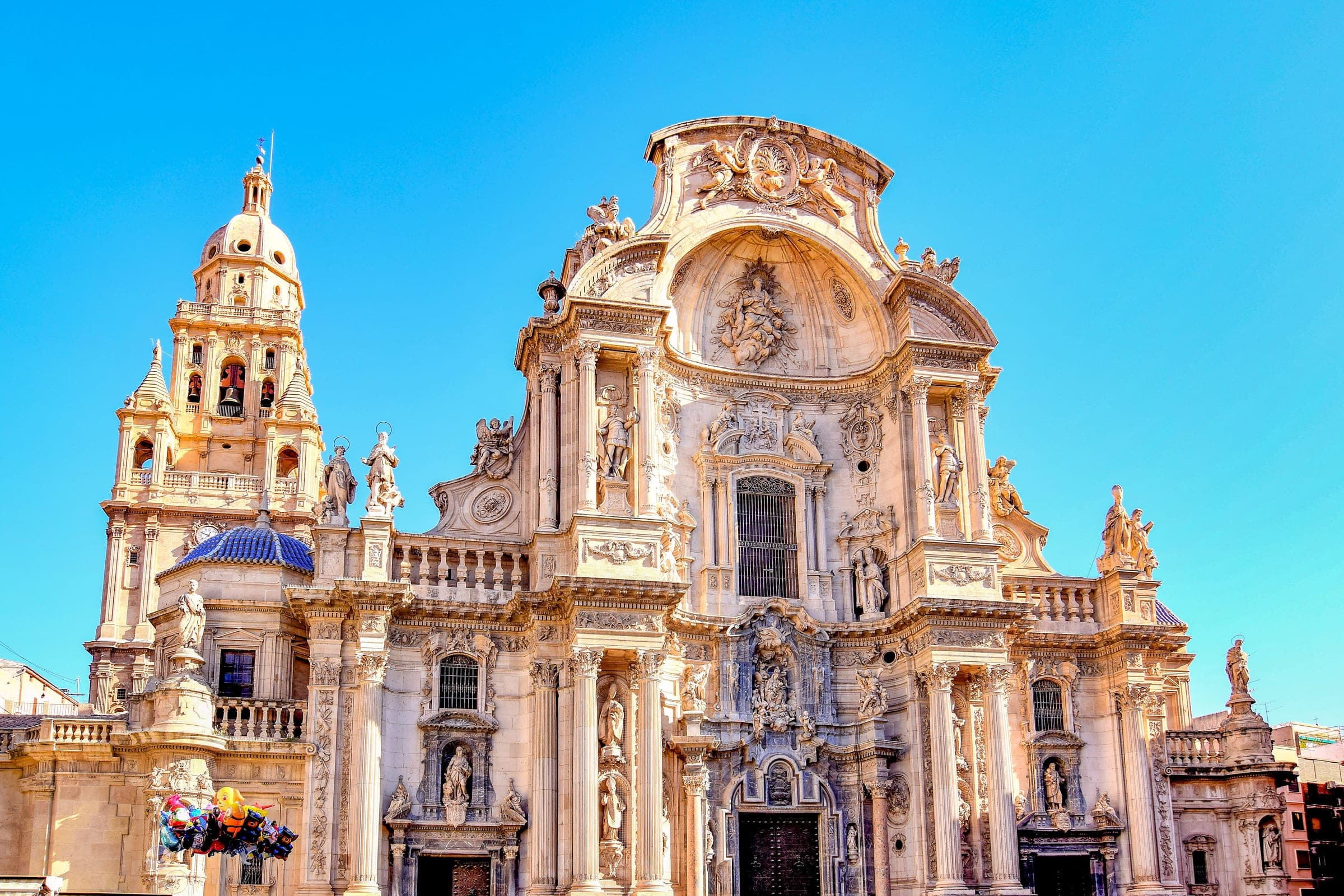 The Baroque west front of Murcia Cathedral on Plaza Cardenal Belluga, with carved saints in niches and the domed bell tower on the left, the square where the Maratón de Murcia finishes