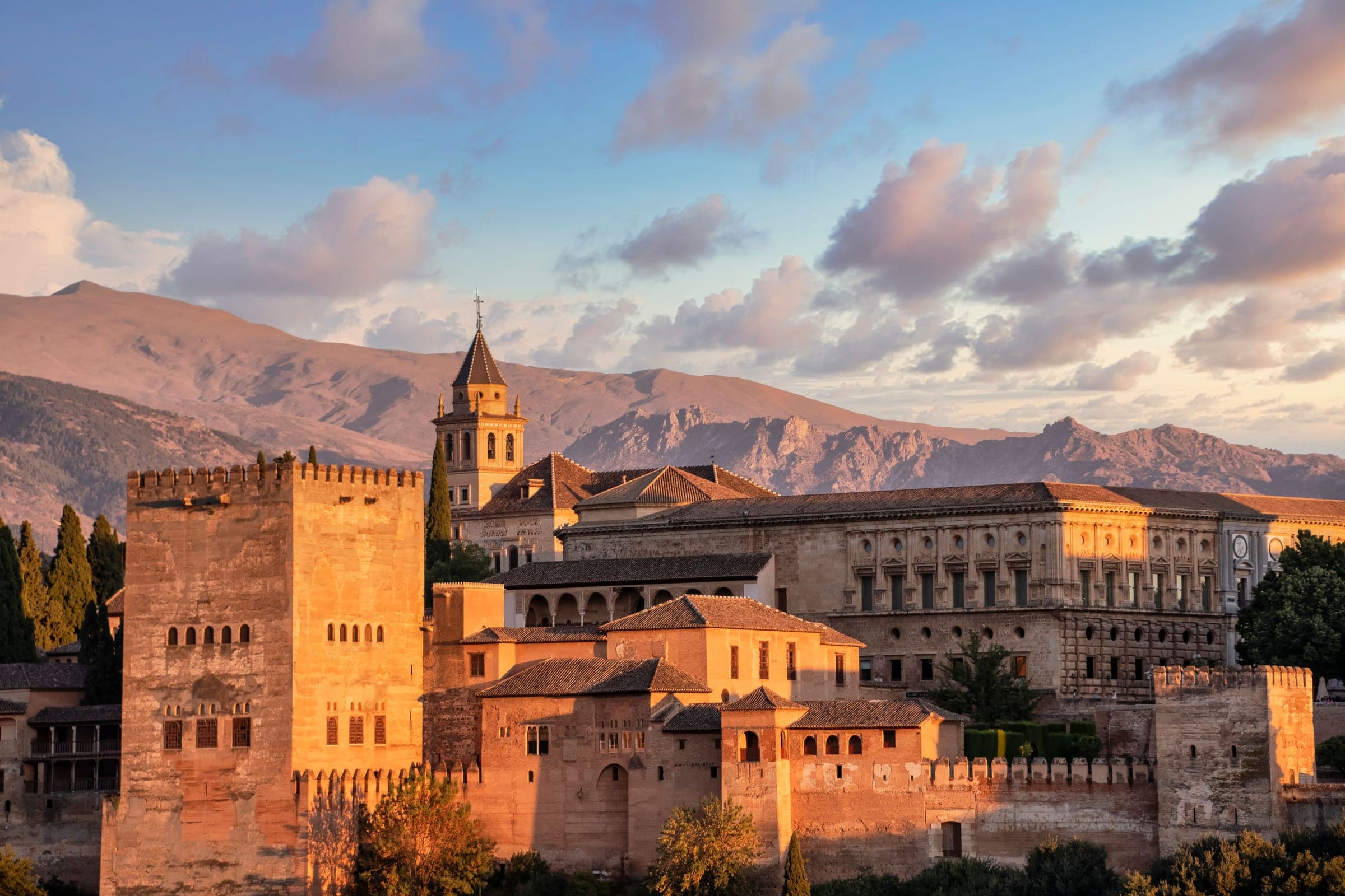 The Alhambra’s towers and the Palace of Charles V glowing in low sun on their hill above Granada, with the bare ridges of the Sierra Nevada foothills behind