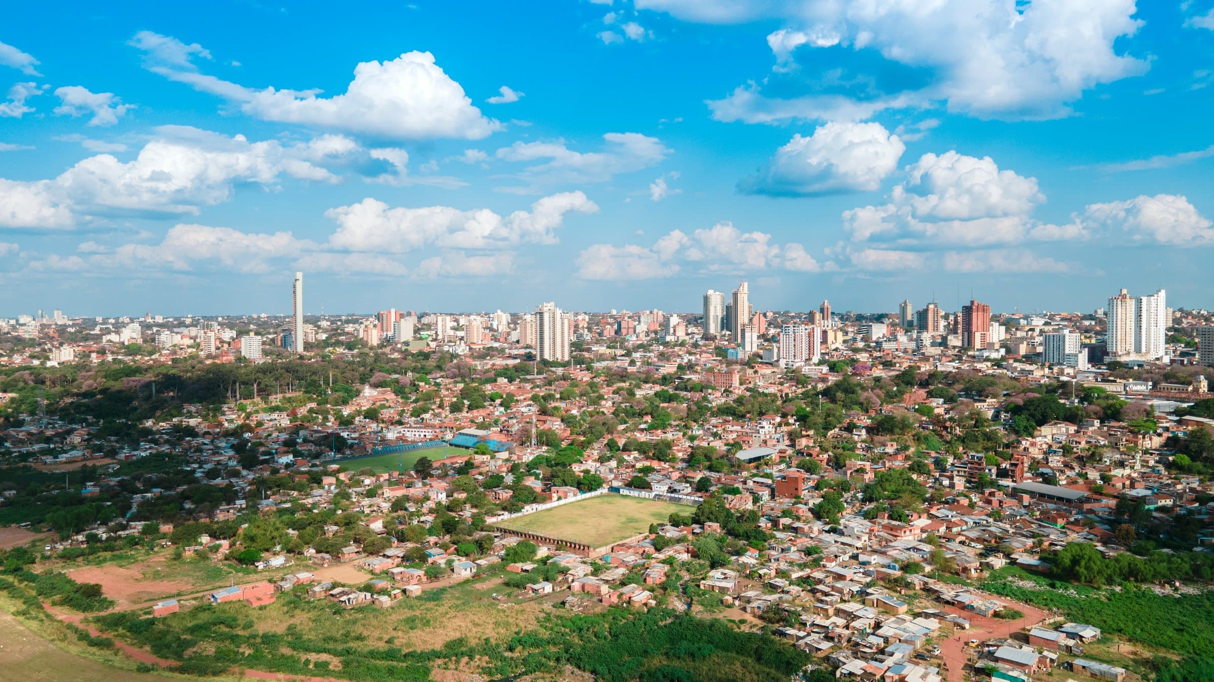 Aerial view over the red-roofed neighbourhoods of Asunción toward the downtown towers on the skyline, under a blue sky with scattered cumulus clouds