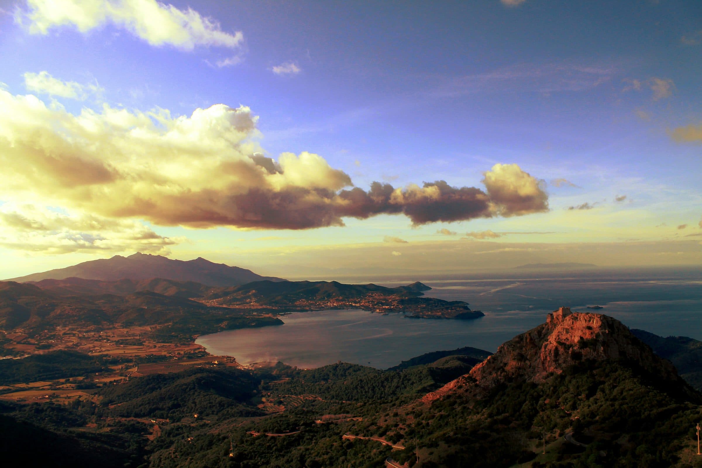 A view over Elba at dusk, with Monte Capanne on the left, a wide bay below and a rocky hill in the foreground under golden clouds