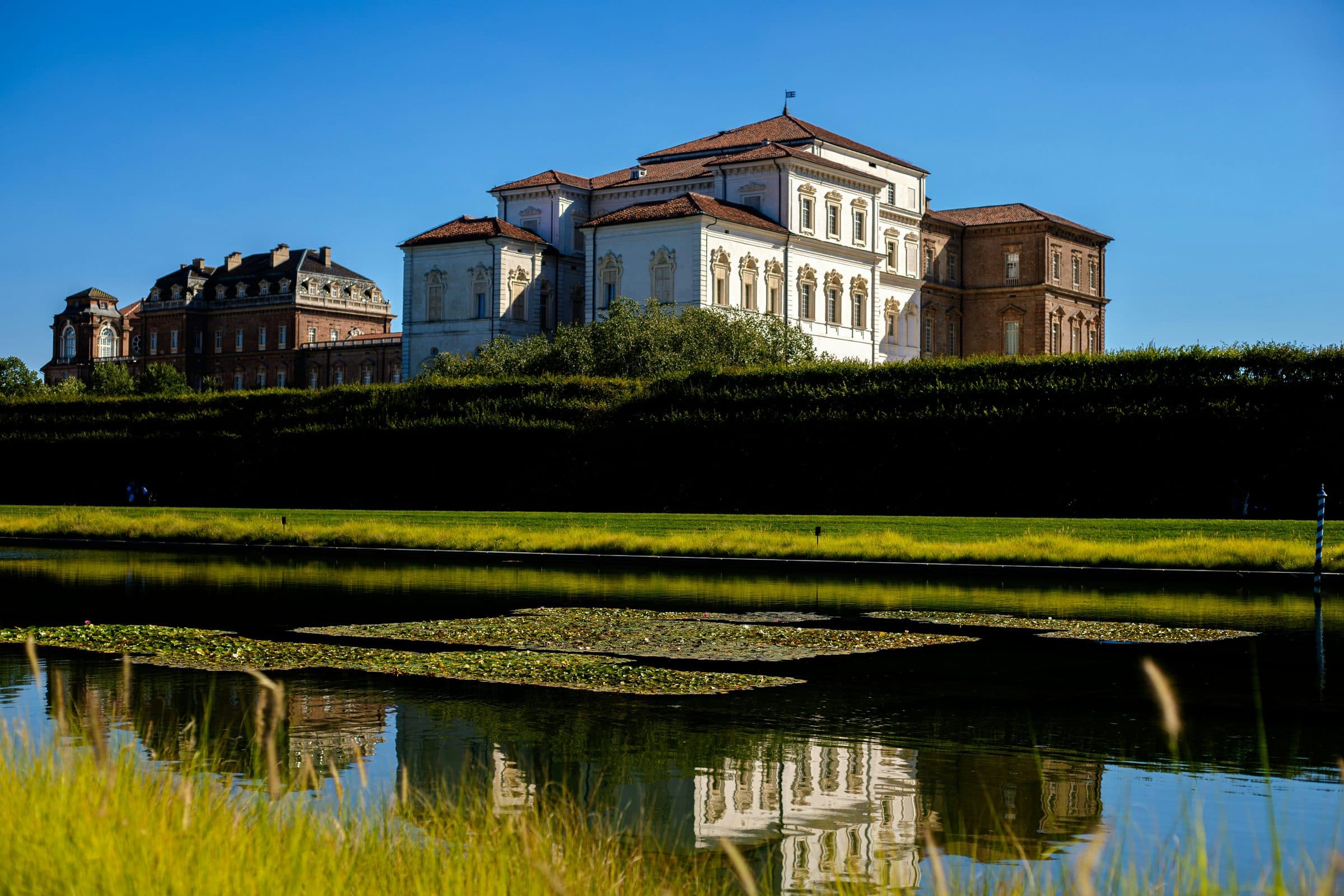 The Reggia di Venaria, a white and brick Savoy palace, seen across a lily pond in its gardens under a clear blue sky