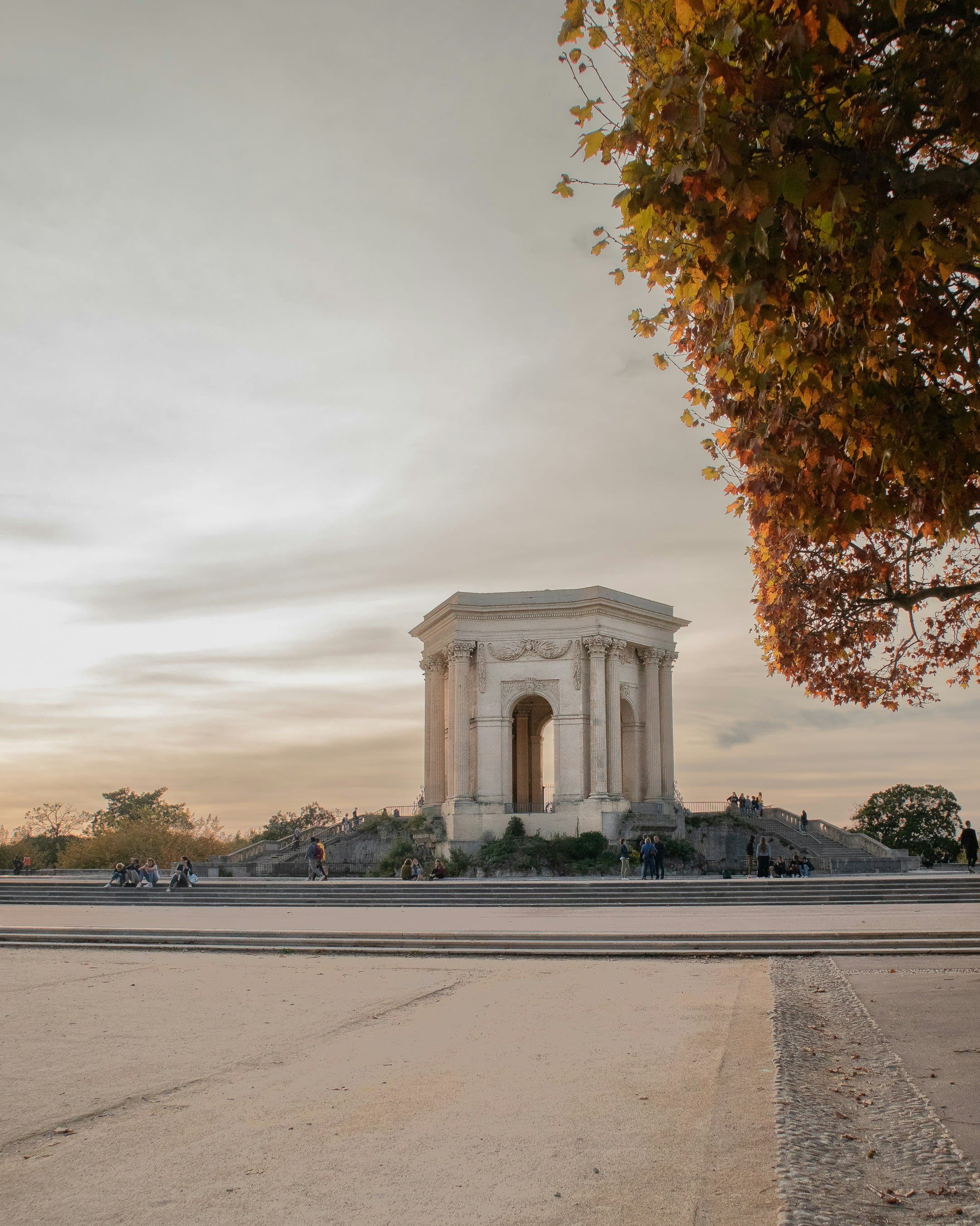 The Château d'Eau, an 18th-century hexagonal water tower with Corinthian columns, at the end of the Promenade du Peyrou in Montpellier, framed by autumn plane-tree leaves at dusk; the race village and bag drop are on this promenade