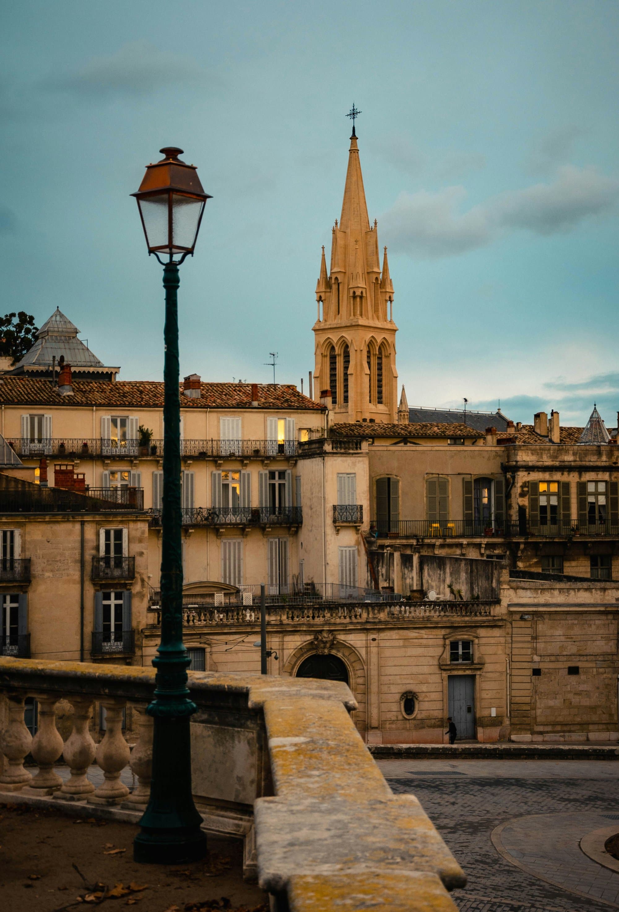 The Gothic spire of the Sainte-Anne church rising above sandstone houses in Montpellier's Écusson, seen past a stone balustrade and an old street lamp at dusk