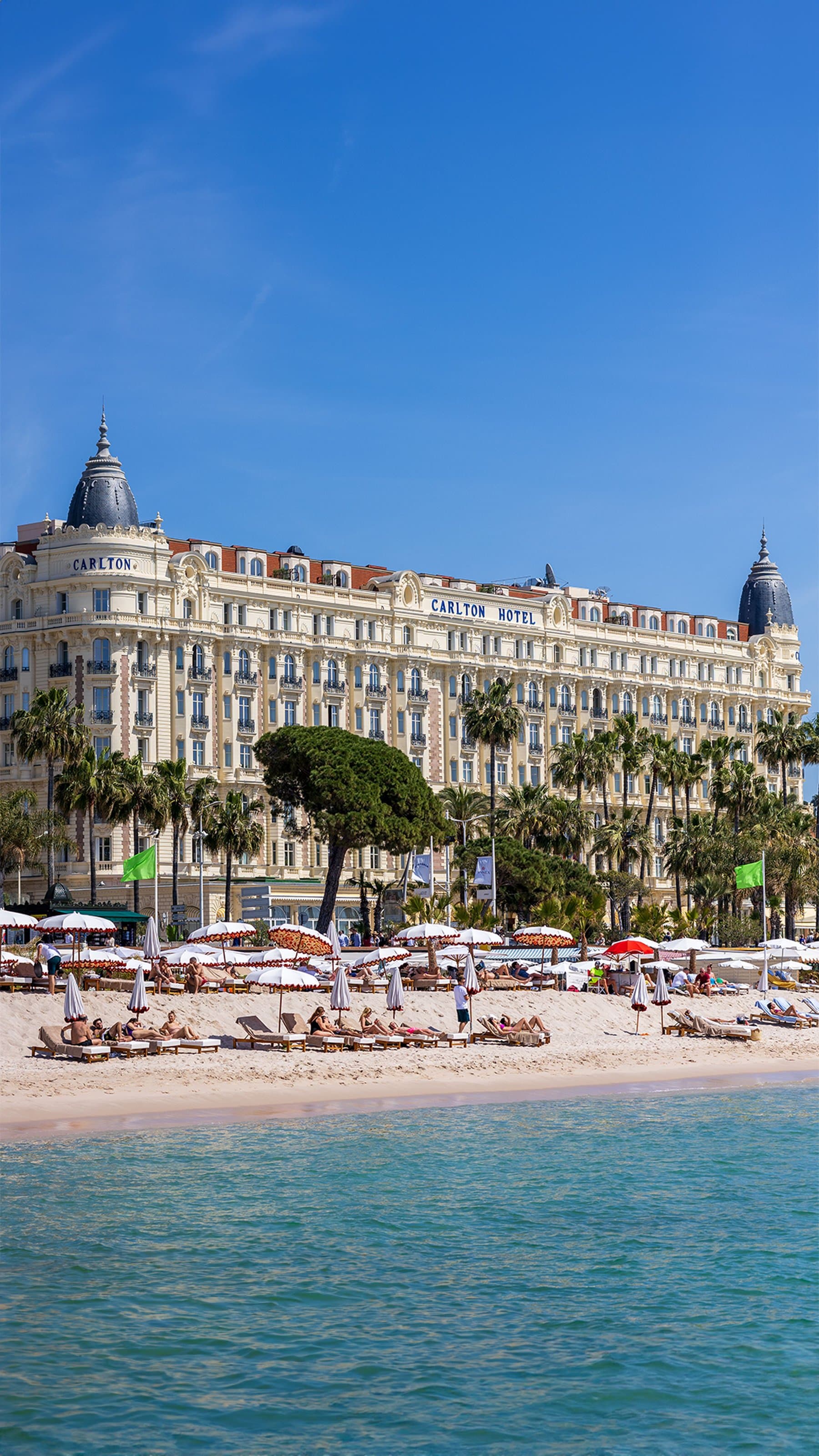 The Carlton Hotel on the Croisette in Cannes, a white Belle Époque facade with two dark domes, behind palm trees and a beach of parasols; the marathon’s last kilometres run along this boulevard
