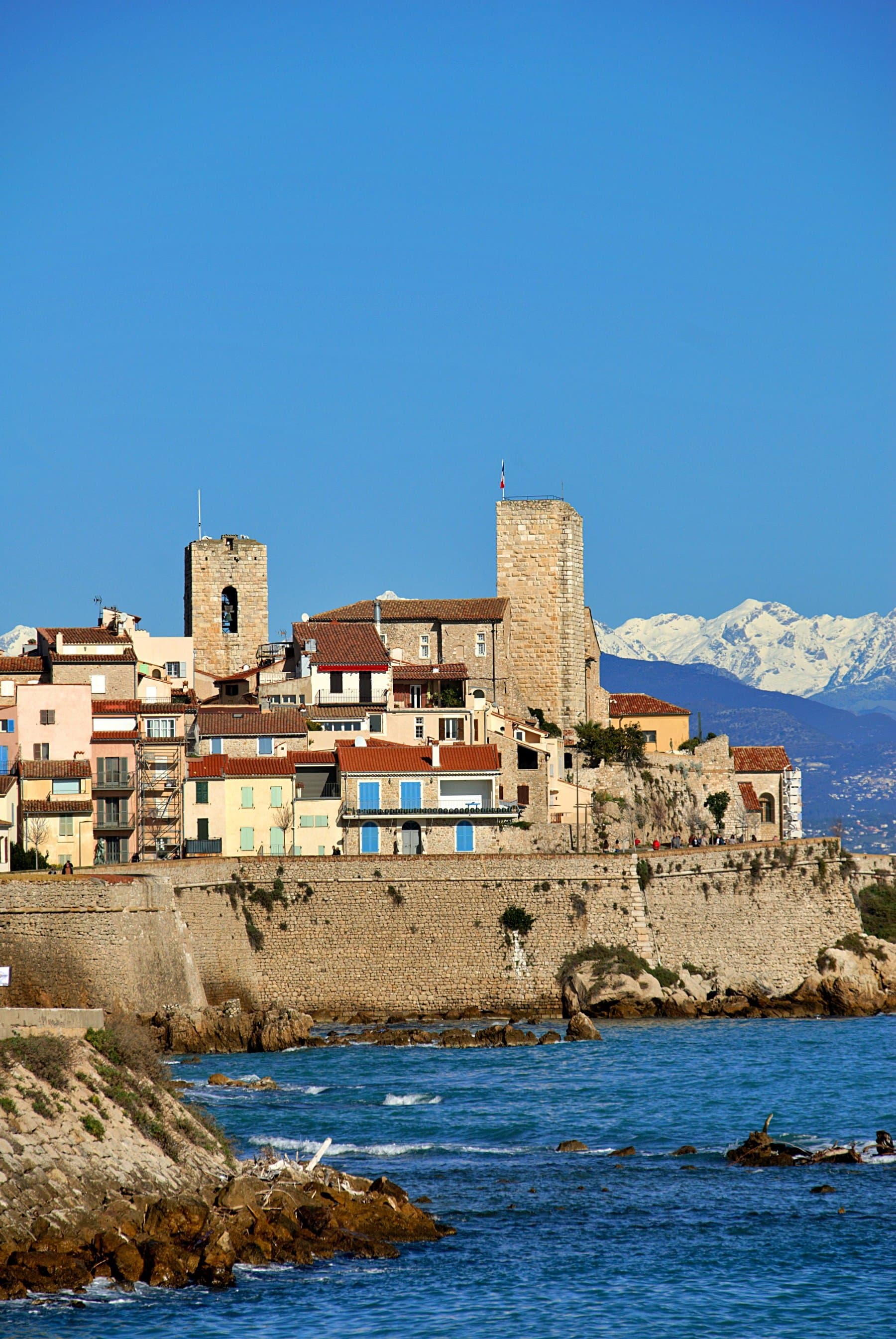 The old town of Antibes on its sea wall, with two stone towers and pastel houses above rocks, and the snow-covered Alps behind; the course runs along these ramparts at km 23