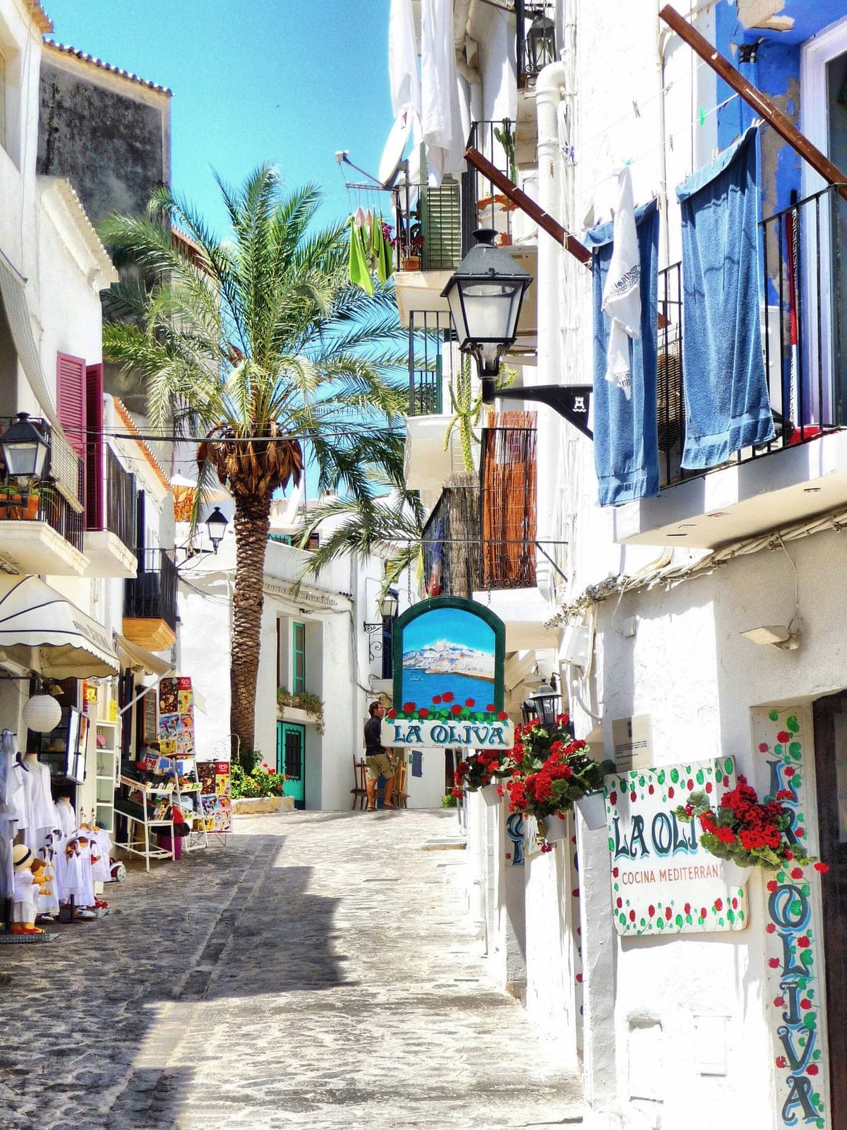 A whitewashed lane in Ibiza’s old town with a palm tree, hanging lanterns and a painted restaurant sign, a short walk up from the marathon start in the port