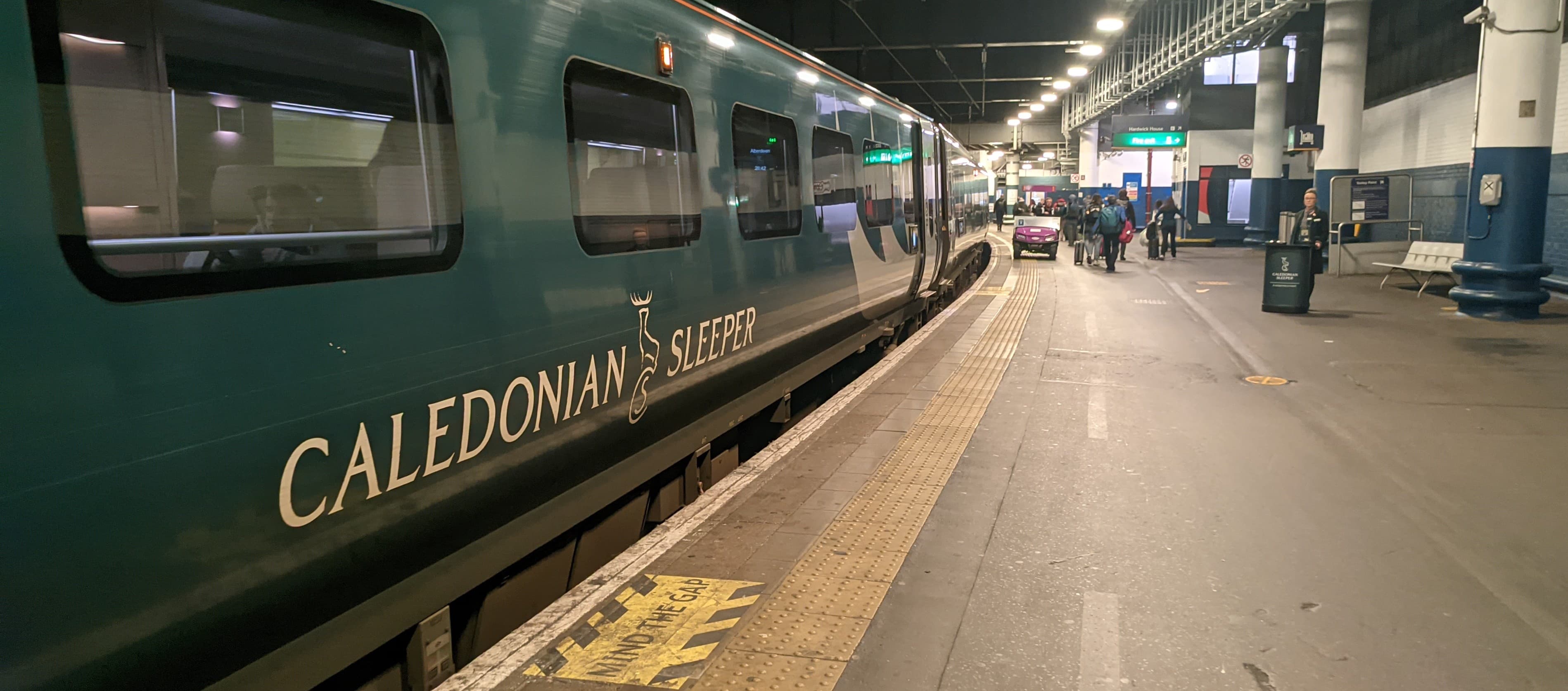 The Caledonian Sleeper train in a Scottish Highland station at dusk, carriages illuminated against a blue-grey sky