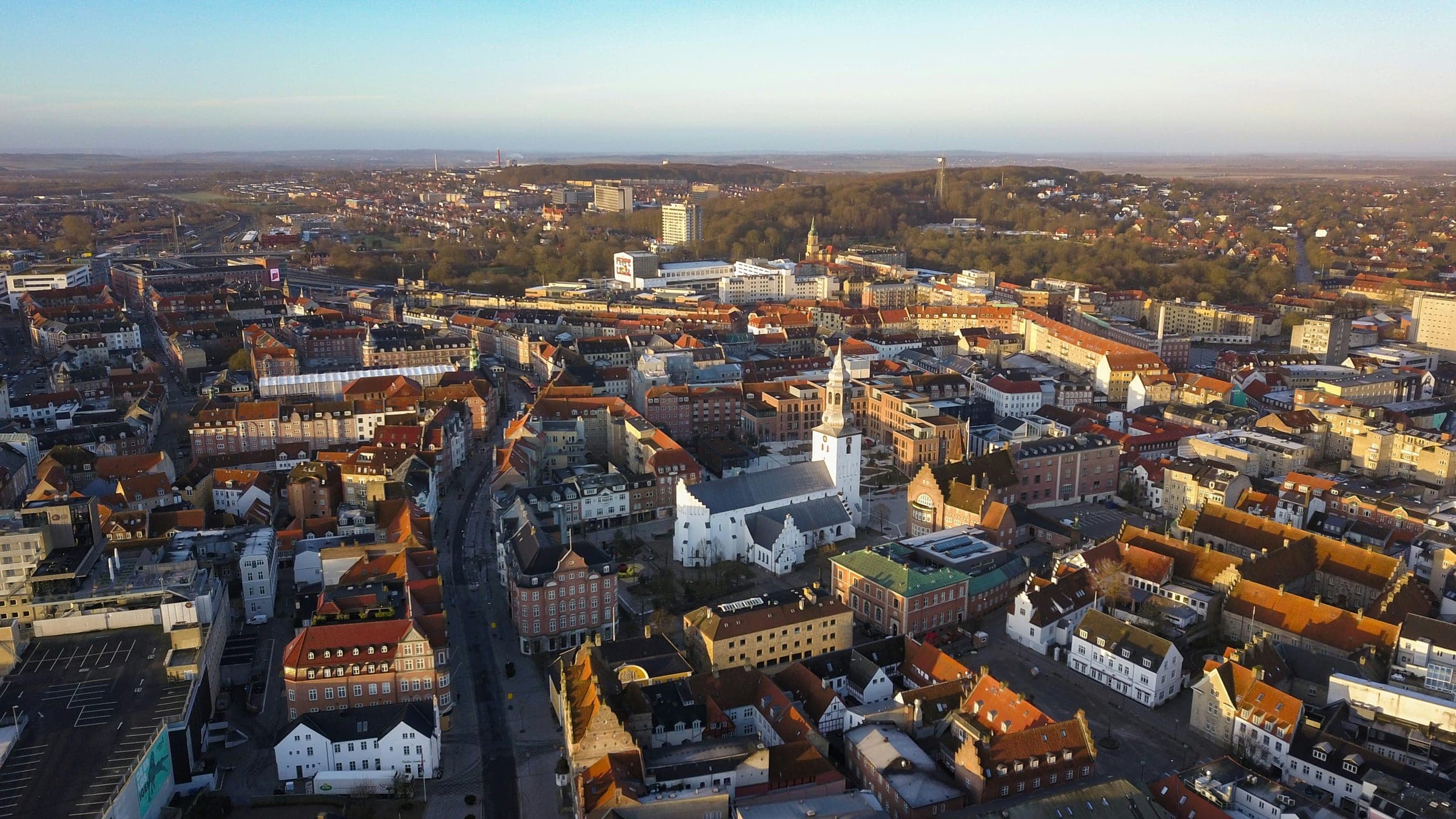 Aerial view of central Aalborg in morning light, with the white tower of Budolfi Church among red-tiled roofs and wooded hills behind the city