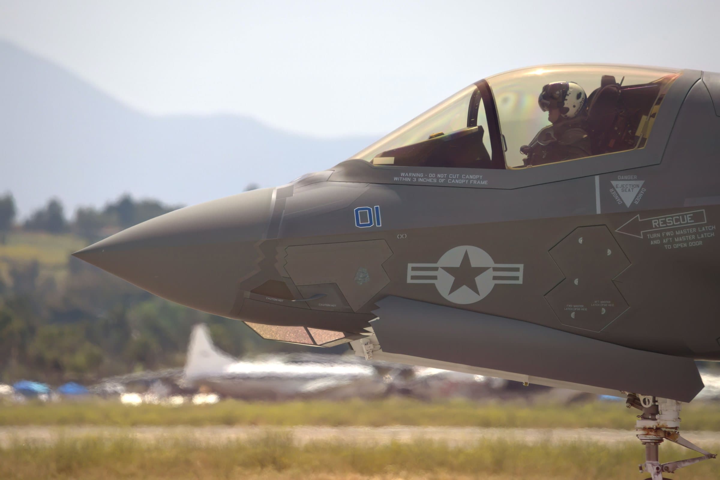 Nose and cockpit of a grey fighter jet on an airfield ramp, the pilot visible under the canopy, with hazy hills and parked aircraft behind