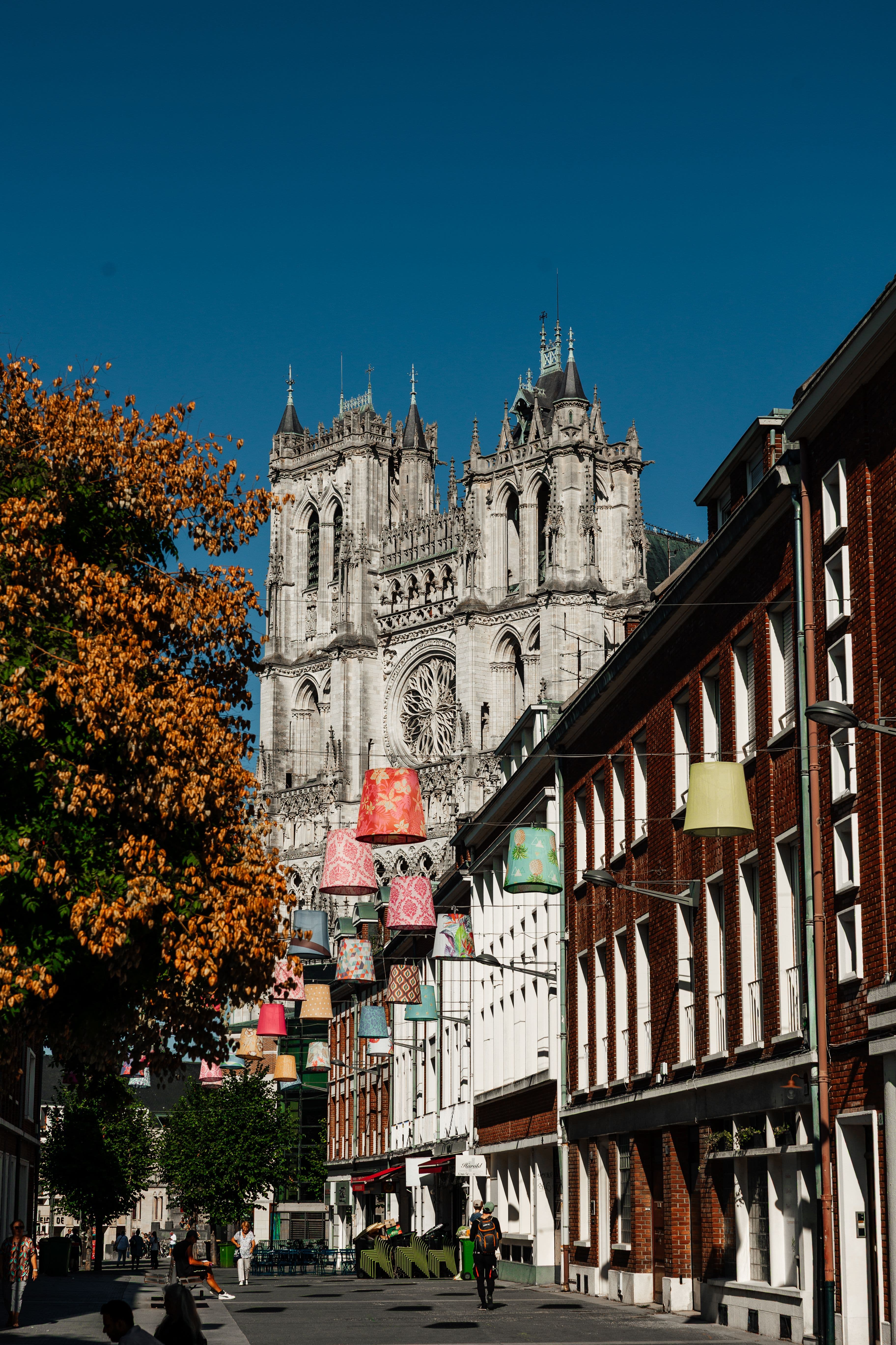The west front of Amiens Cathedral at the end of a narrow street hung with coloured fabric lampshades, under a clear blue sky, with an autumn-coloured tree in the foreground.