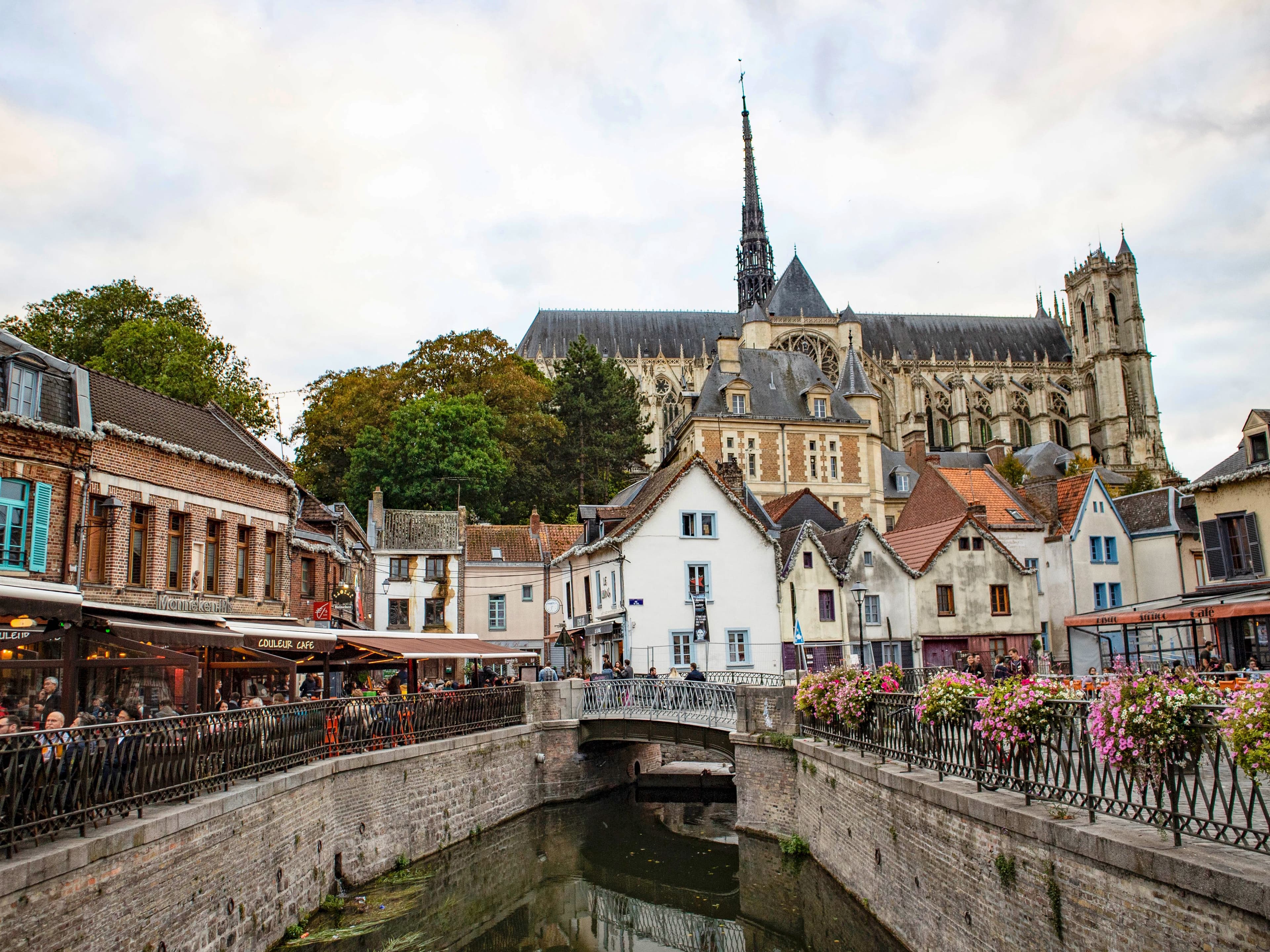 Amiens Cathedral rising above the Saint-Leu quarter, seen from a small canal bridge lined with flower boxes, with café terraces along the water. The marathon starts about 4 km north-west of here, at Parc du Grand Marais.