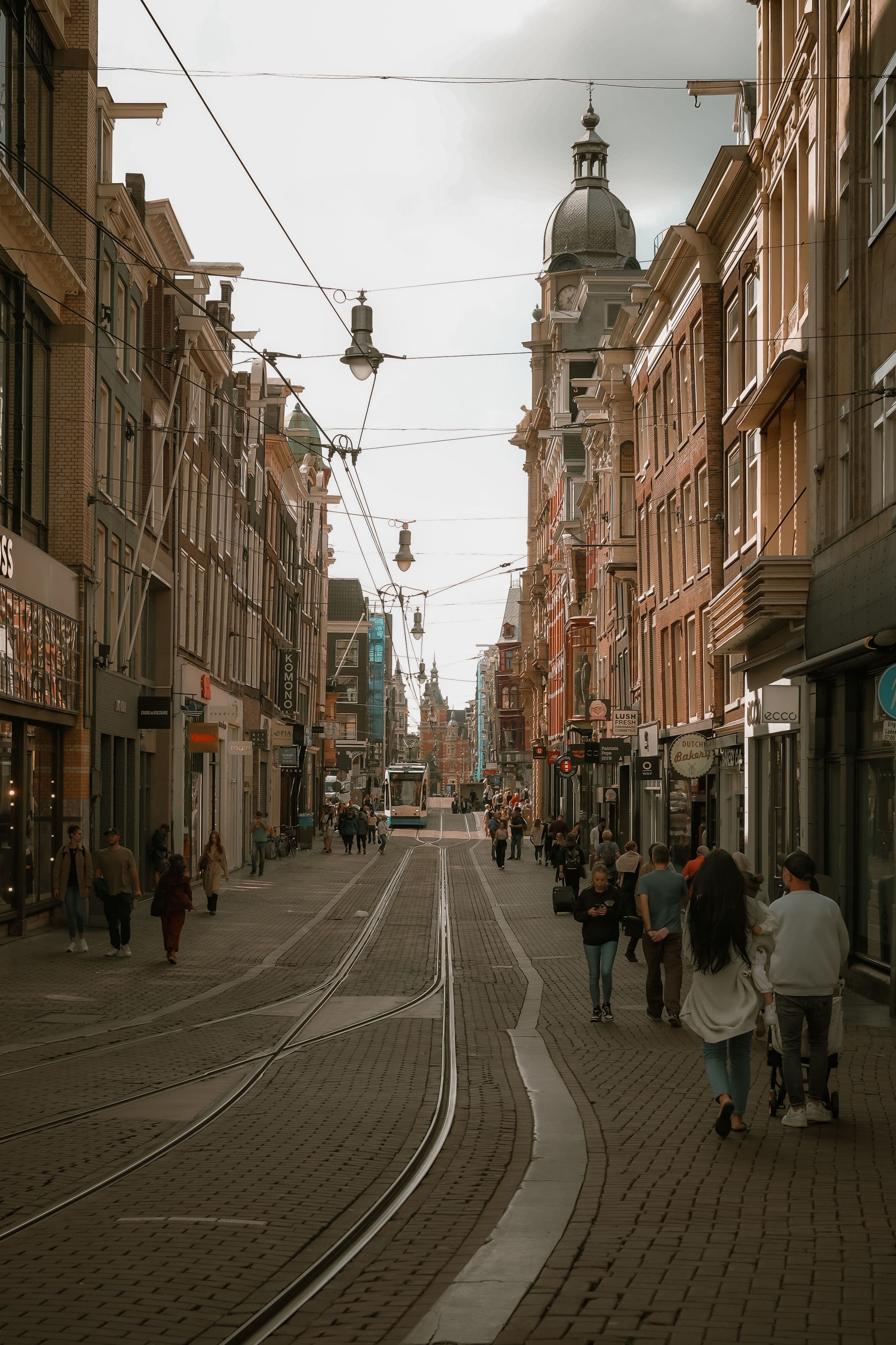 A tram running down a central Amsterdam shopping street lined with narrow gabled buildings and overhead tram wires
