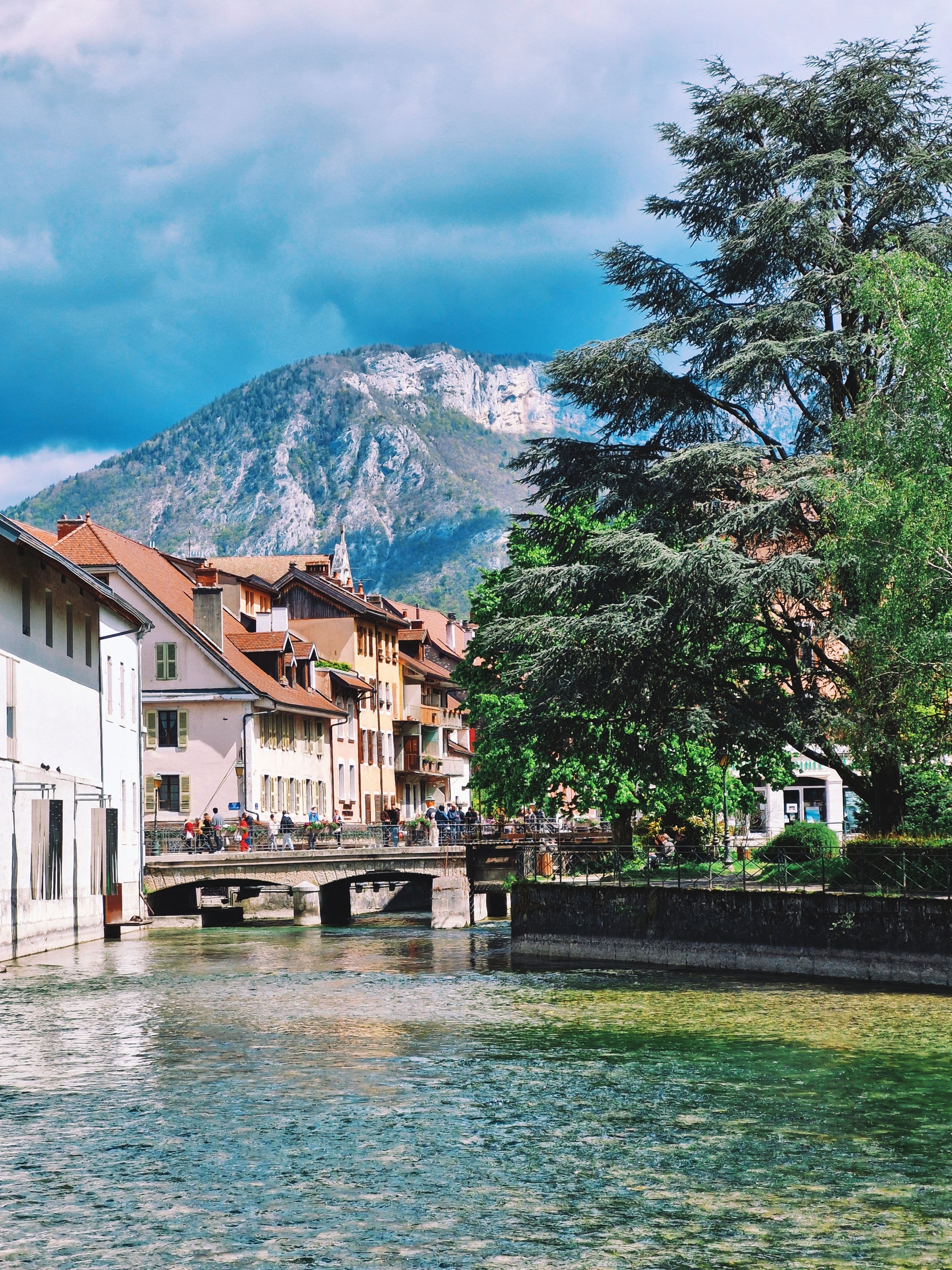 The clear green water of the Thiou canal in Annecy's old town, with a stone footbridge, pastel houses and a rocky mountain peak behind under a stormy sky. The old town is a few minutes' walk from the Le Pâquier finish.