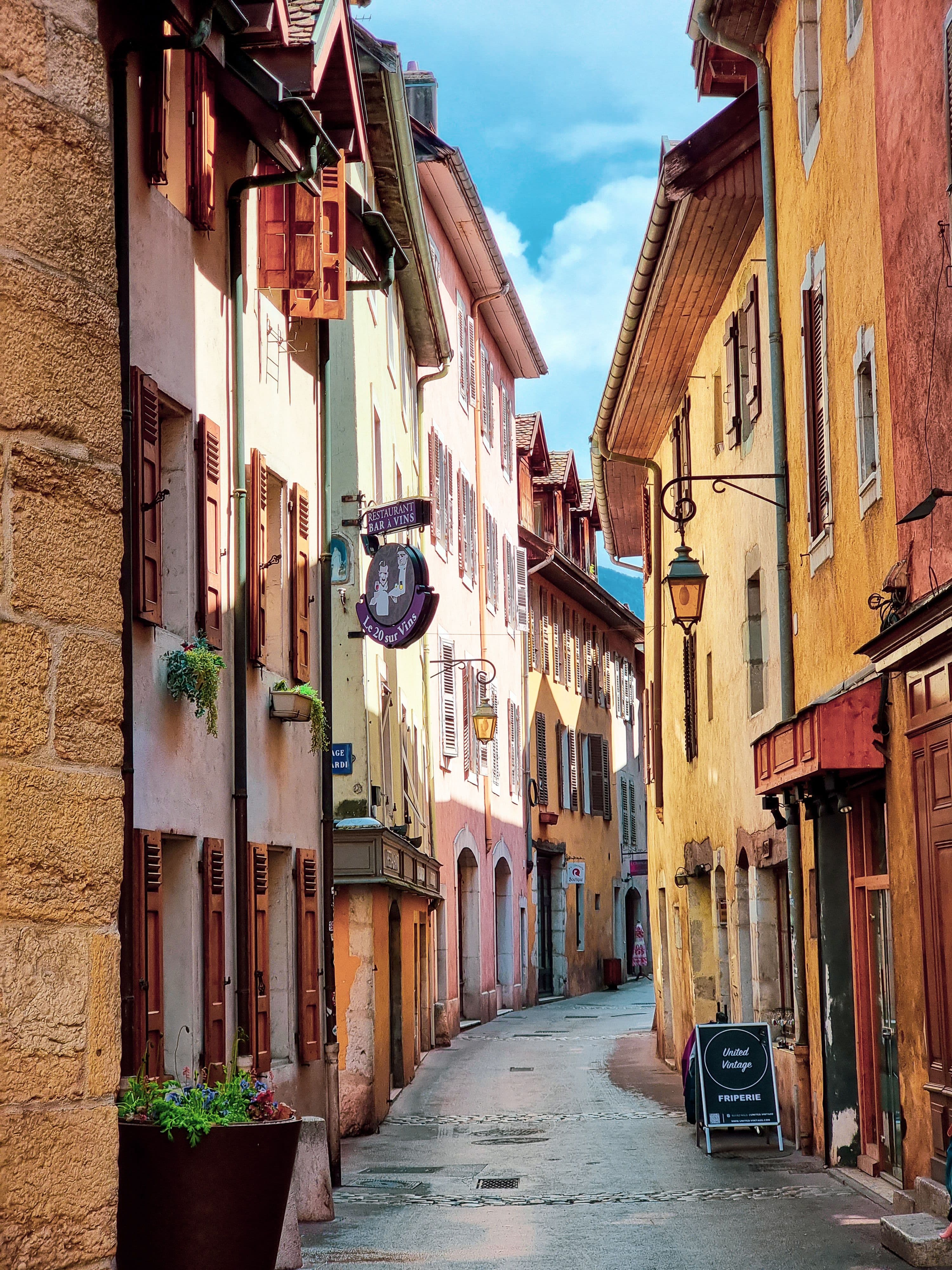 A narrow cobbled street in Annecy's old town lined with ochre and pink houses, wooden shutters, hanging shop signs and iron lanterns.