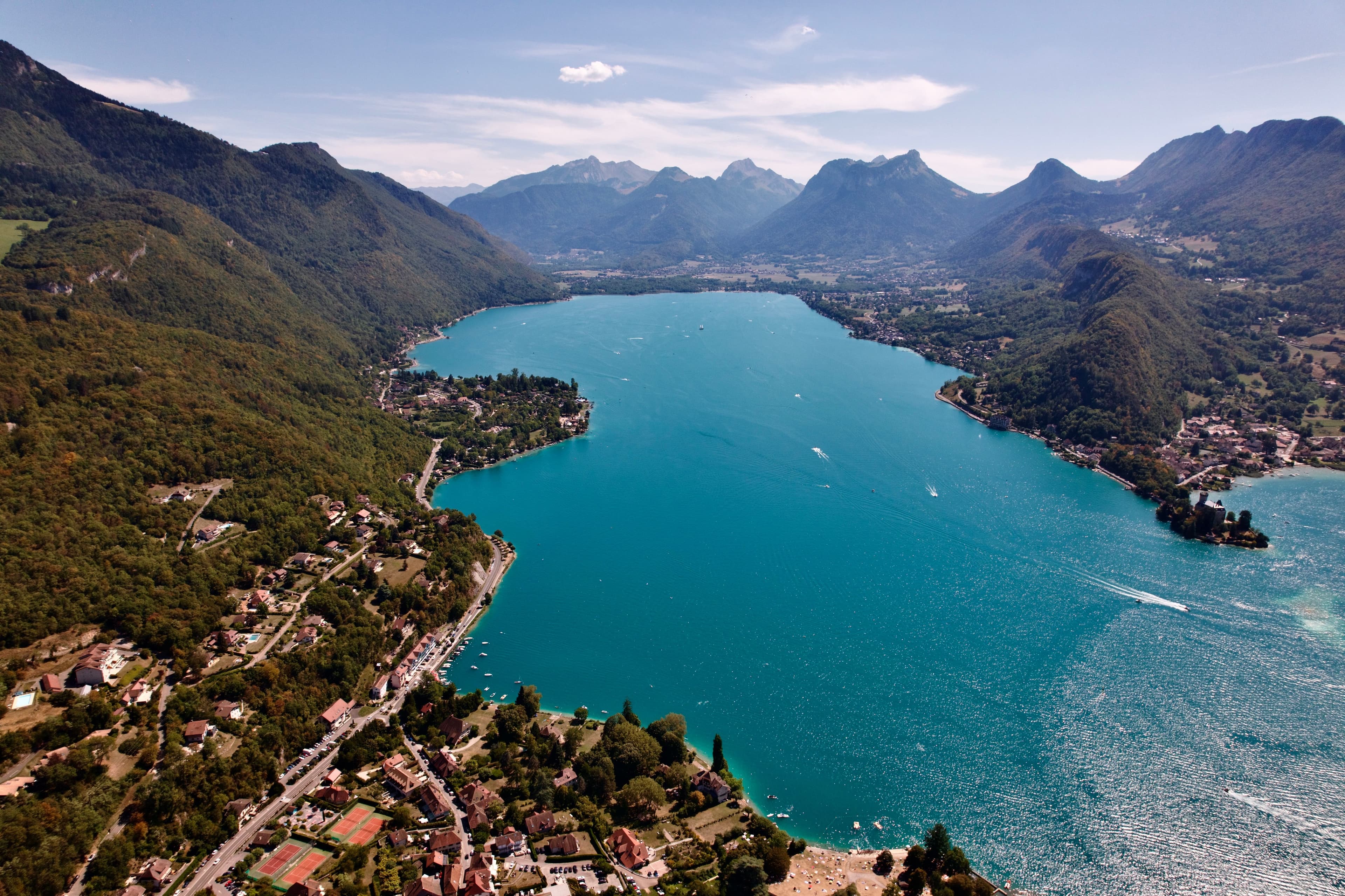 Aerial view of Lake Annecy's turquoise water between steep wooded mountains, with villages along both shores and a small castle on a spit of land. The marathon runs along the western shore to the southern end of the lake and back.