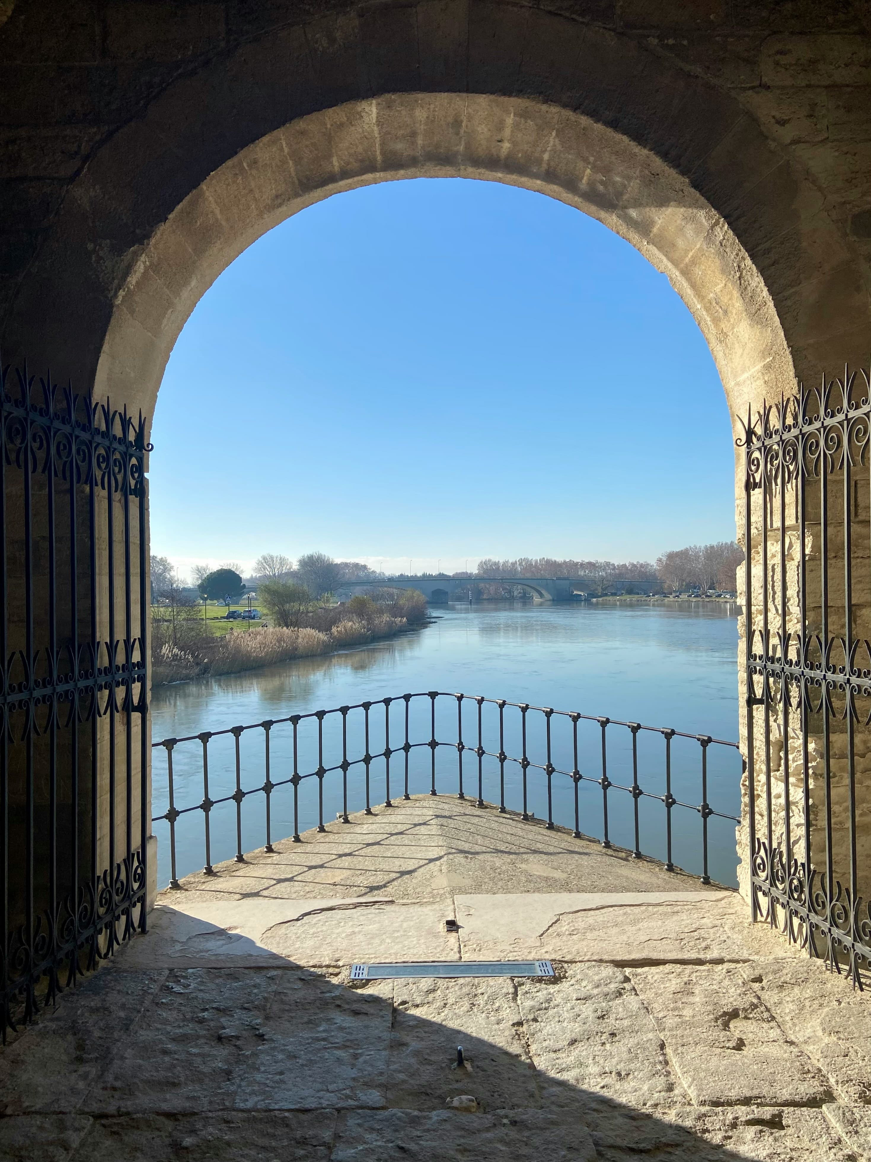 The Rhône seen through a stone arch in Avignon's riverside wall, wrought-iron gates open on either side, with the modern Pont Édouard Daladier crossing to Île de la Barthelasse in the distance. The marathon crosses to the island and finishes there.