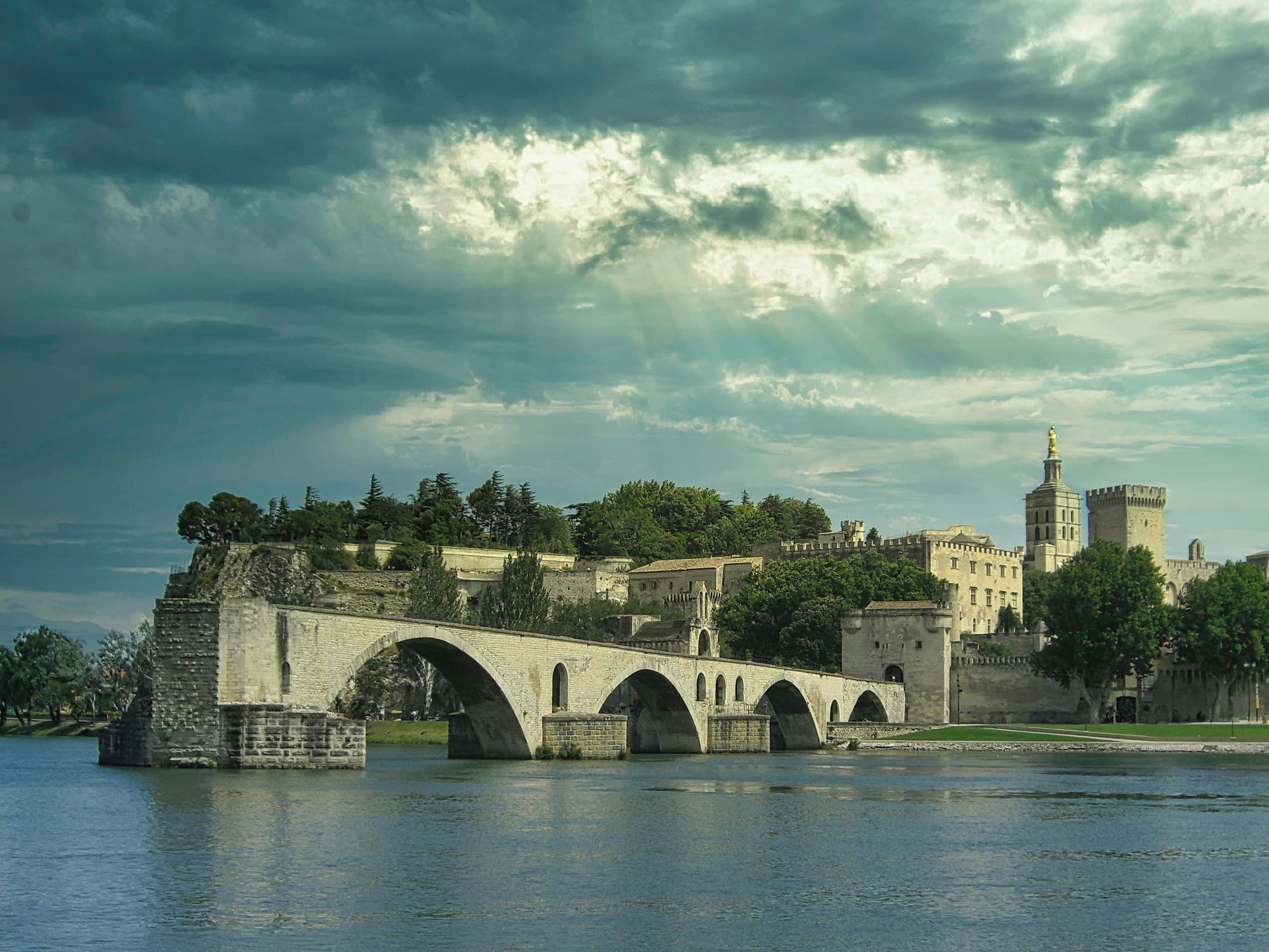 The medieval Pont Saint-Bénézet reaching out over the Rhône at Avignon, with the ramparts, the Palais des Papes and the cathedral's gilded statue on the skyline behind it under a broken sky. The marathon passes this stretch of riverbank inside its first eight kilometres.