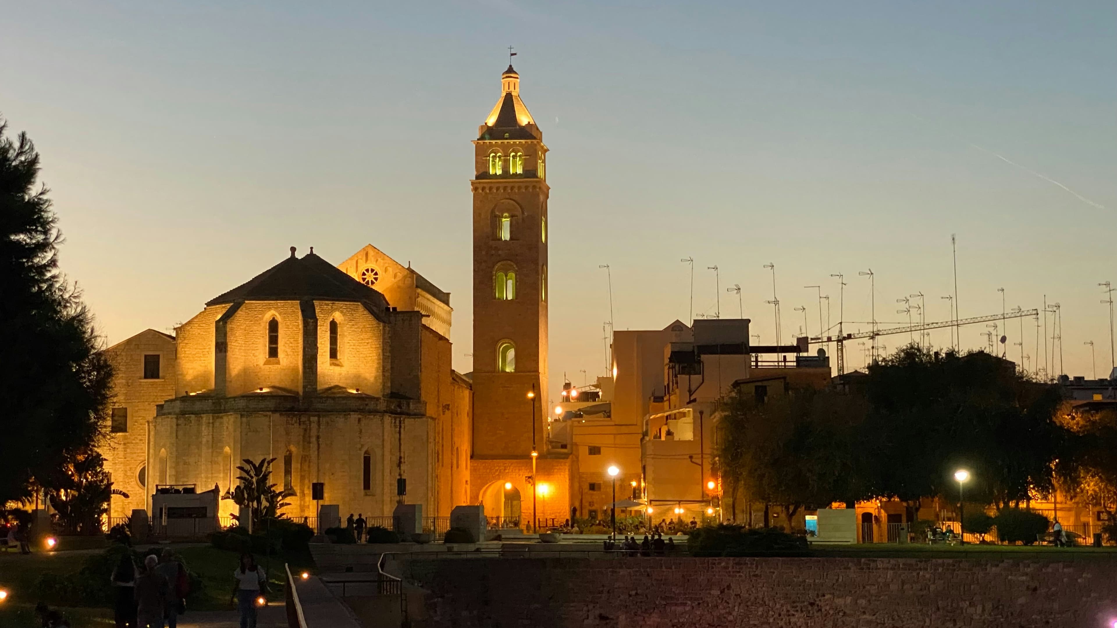 The floodlit apse and brick bell tower of Barletta's cathedral at dusk, seen over the castle moat wall with pines and a park in the foreground