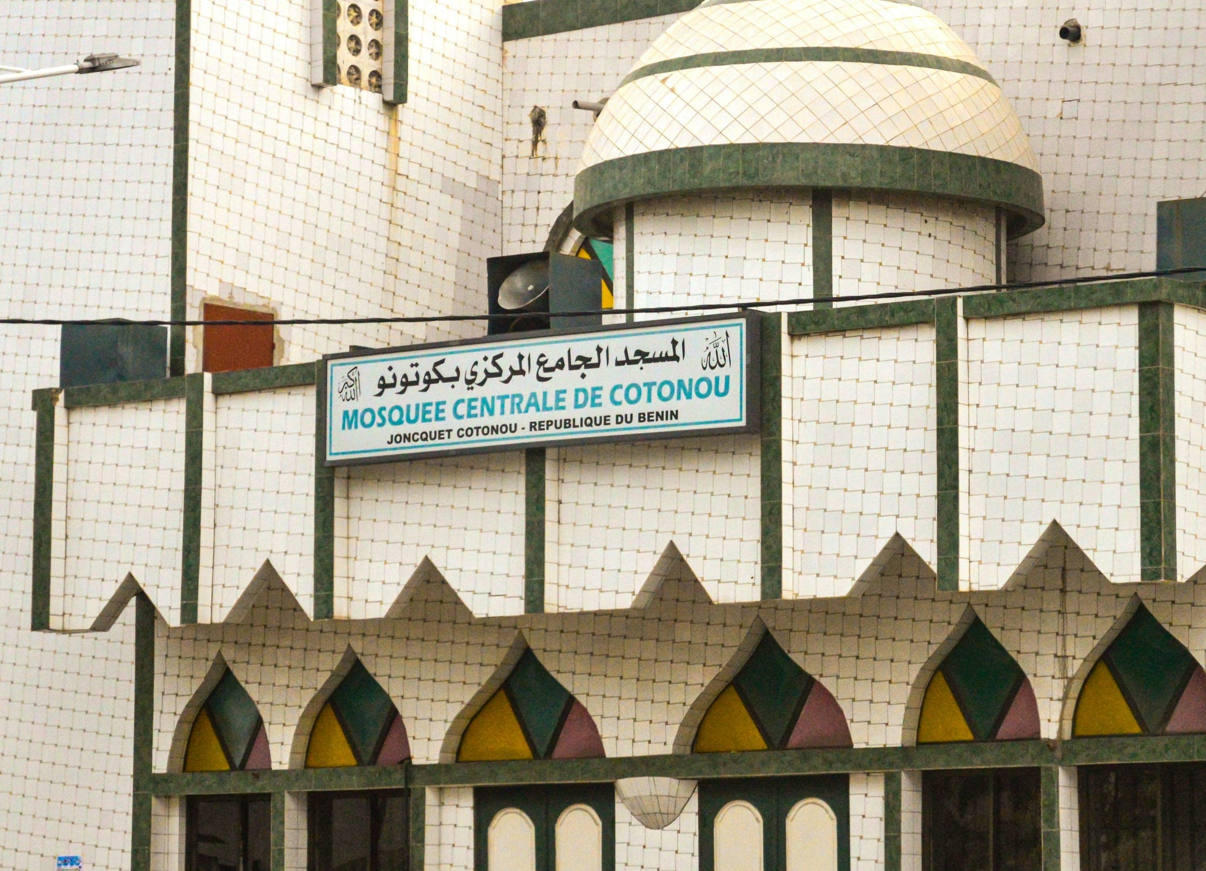 The tiled facade of the Central Mosque of Cotonou, Benin, with a green and white dome, a blue name sign in French and Arabic, and pointed windows in yellow, green and pink glass.