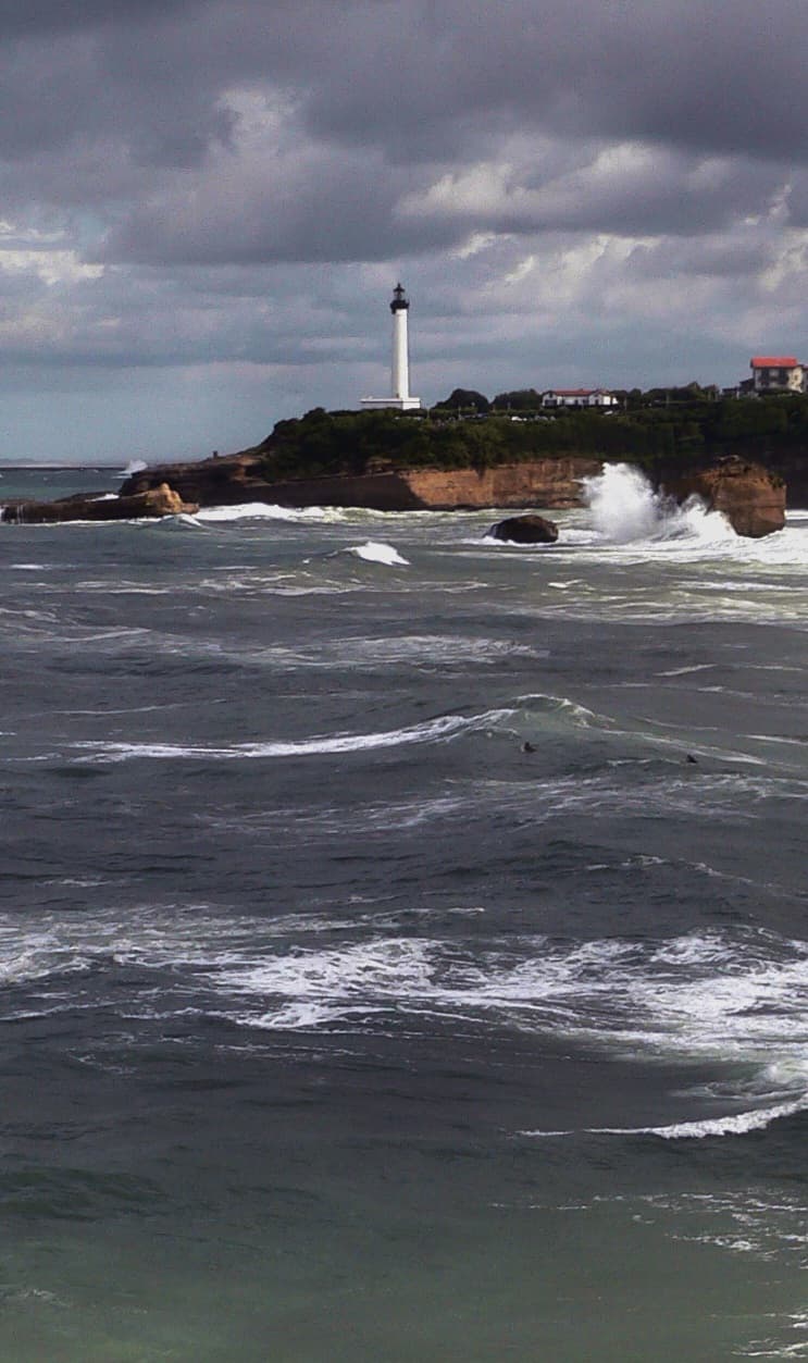 The white Phare de Biarritz lighthouse standing on a green clifftop above the Atlantic, with a heavy swell breaking white against the rocks below under a dark, stormy sky. The lighthouse is one of the landmarks on the course's coastal return.