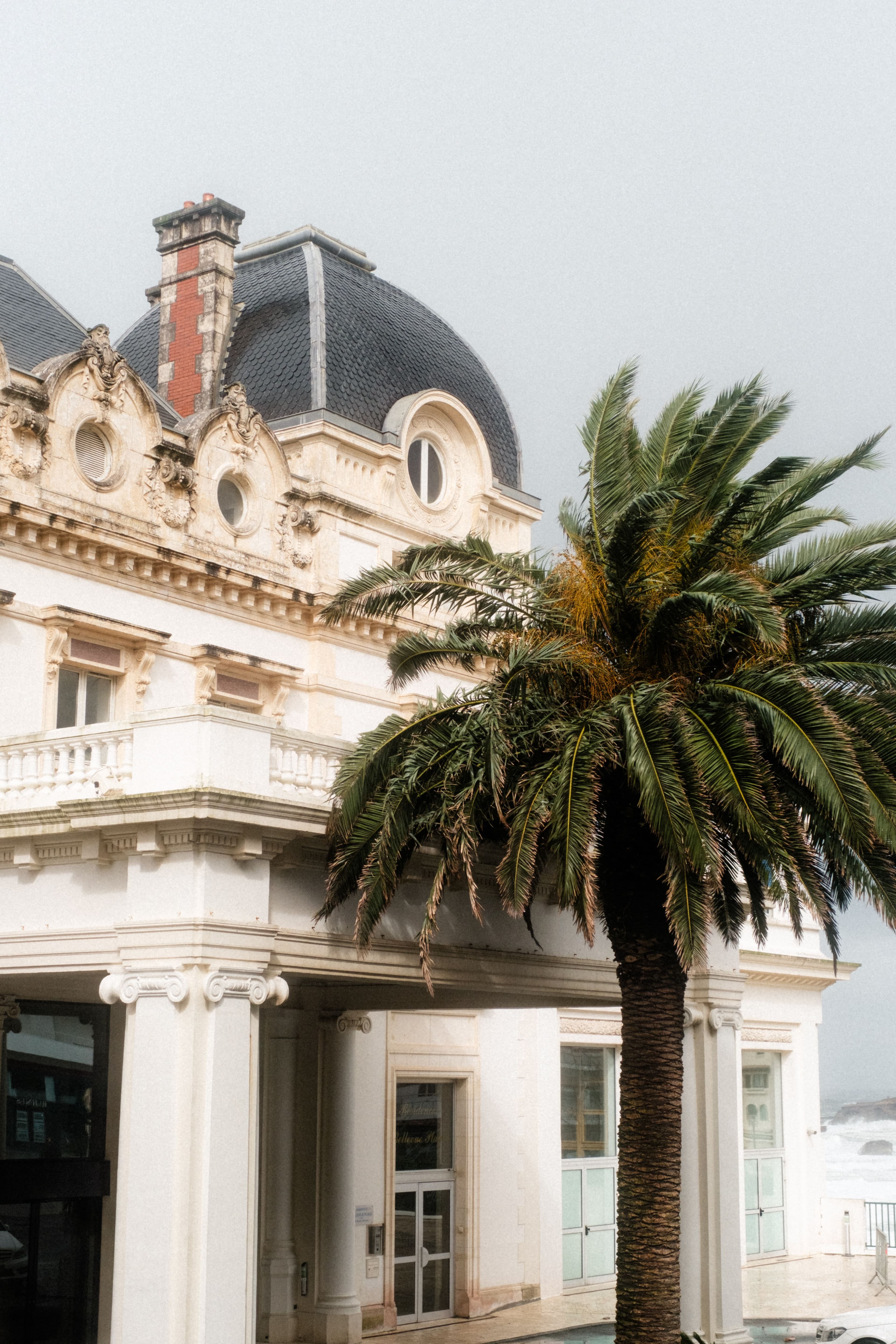 A Belle Époque seafront building in Biarritz with a slate dome, carved stonework and Ionic columns, a wind-blown palm tree in front of it and the grey Atlantic visible to one side.