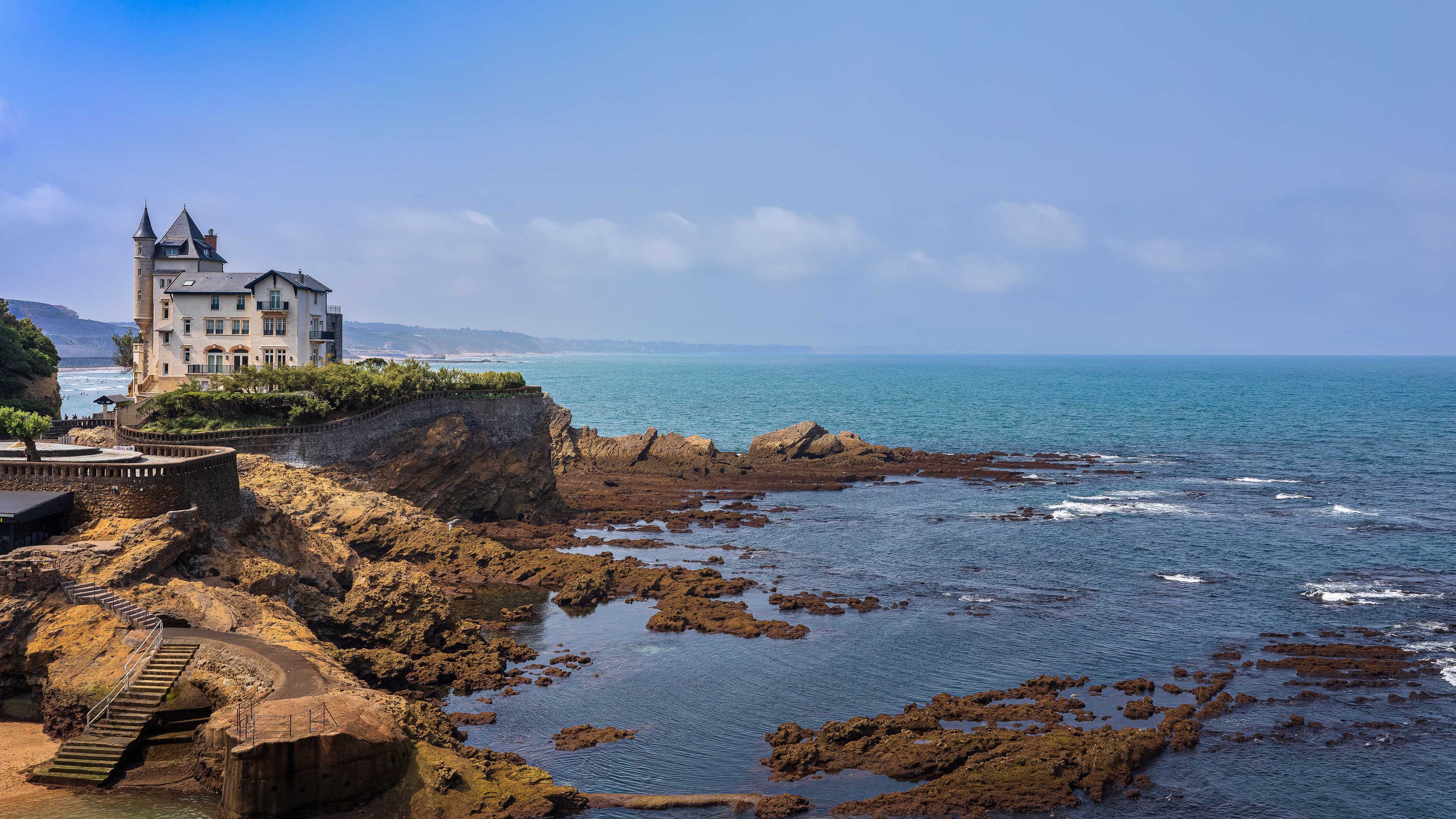 Villa Belza, a turreted stone mansion, standing on a rocky headland above the Atlantic at the Côte des Basques in Biarritz, with seaweed-covered rocks and turquoise water in the foreground. The marathon returns along this coast in its closing kilometres.