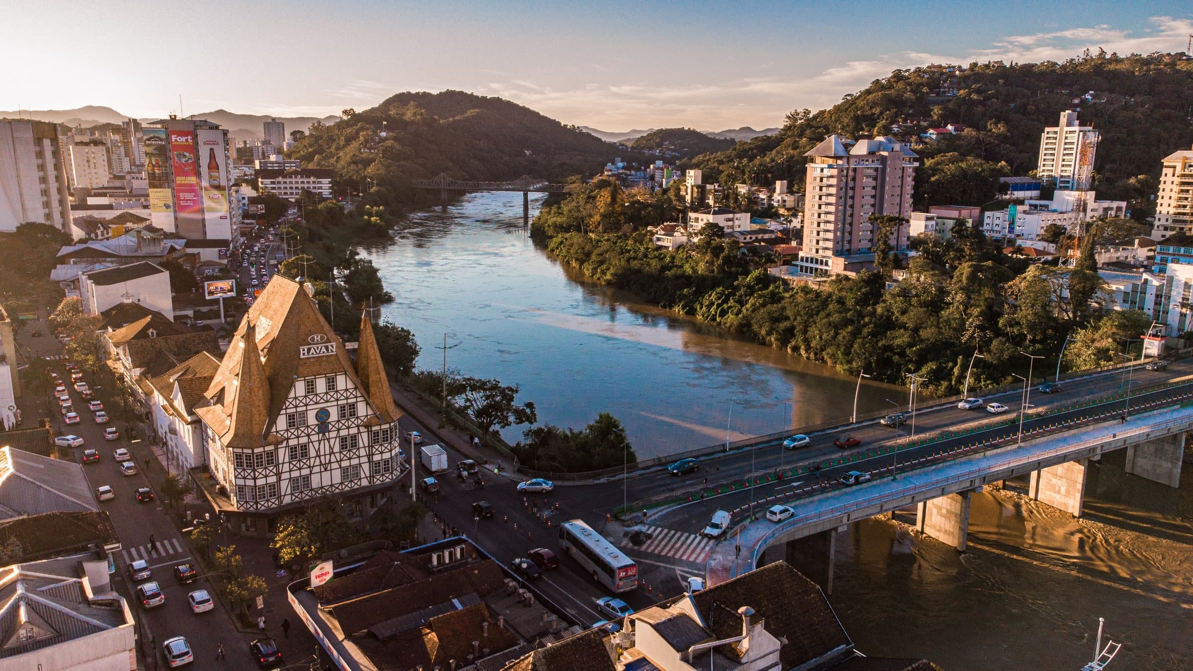 Aerial view of a half-timbered German-style building beside the Itajaí-Açu river in Blumenau, with a road bridge and forested hills in evening light