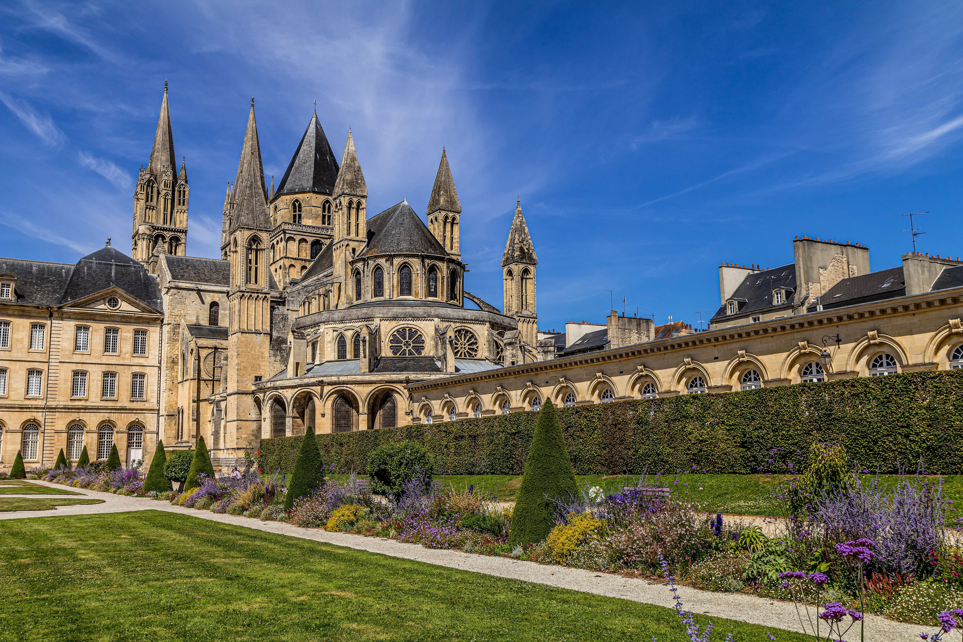 The Abbaye aux Hommes in Caen, its Romanesque church spires and the Hôtel de Ville wing rising above formal gardens and clipped hedges under a blue sky. The marathon's closing kilometres pass the abbey on the way to the stadium finish.