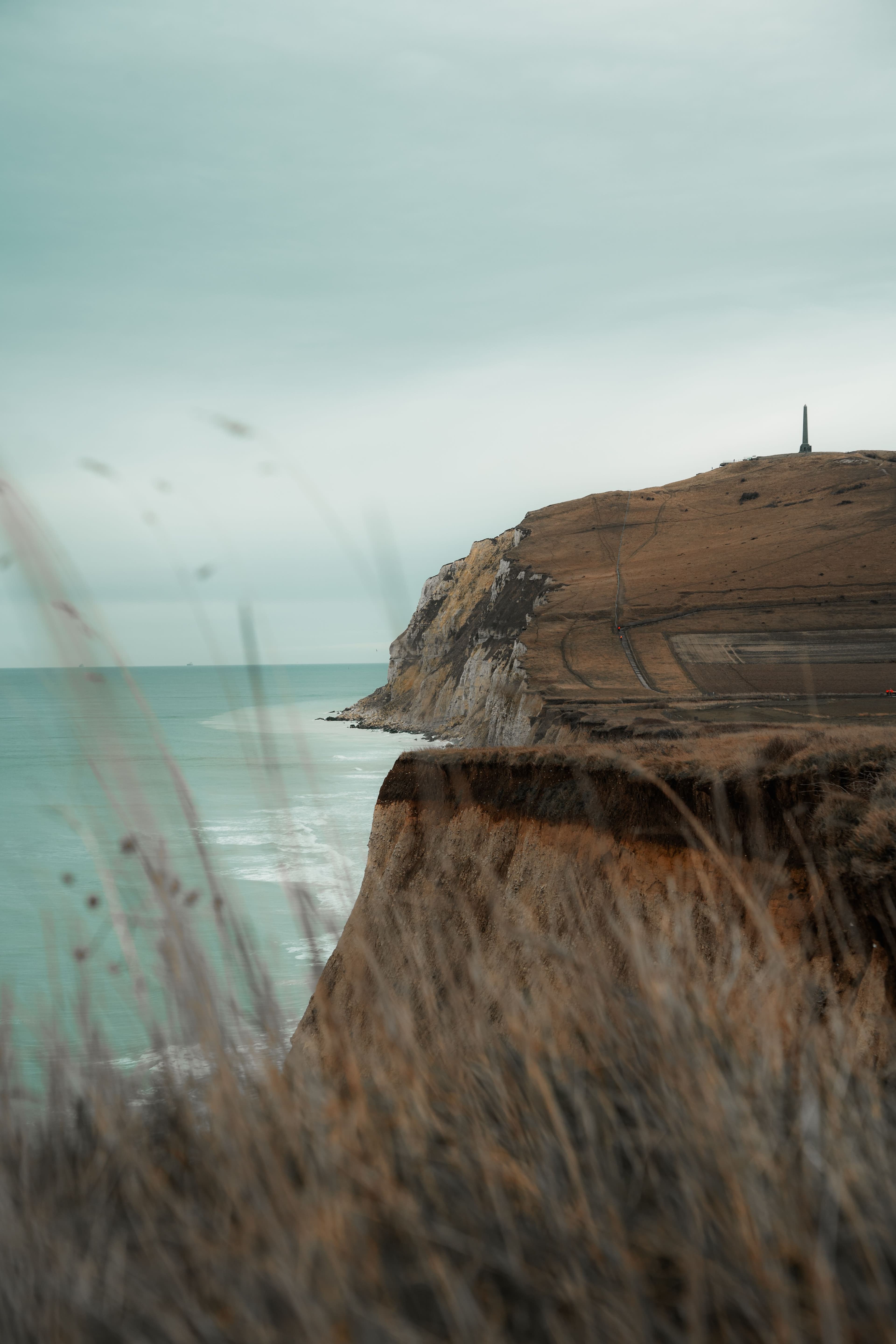 The chalk headland of Cap Blanc-Nez seen along the coast, its white cliff face dropping to the sea with the Dover Patrol obelisk on the summit and dune grass in the foreground. The marathon starts at Escalles beneath it.