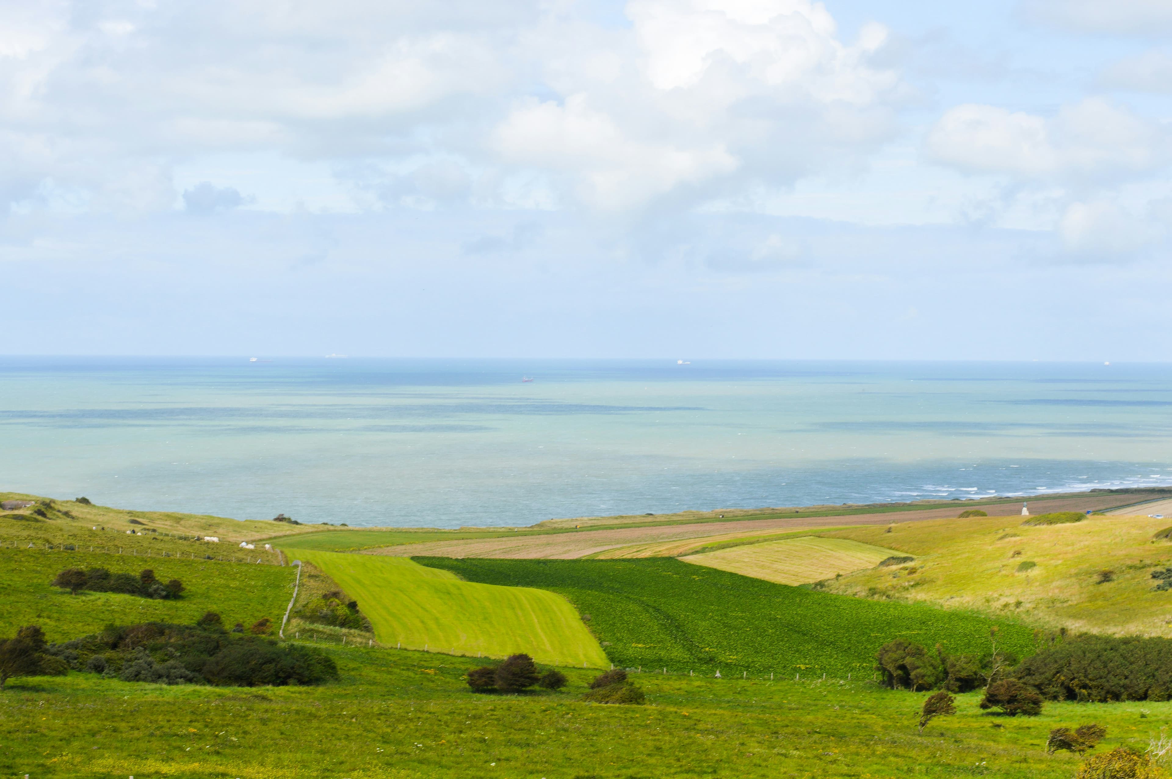 Green farmland and low dunes sloping down to the Strait of Dover on the Opal Coast near Sangatte, with cargo ships on the horizon and the Dover Patrol obelisk on the skyline to the right. The marathon's coast-road section, from Escalles to Sangatte, runs through this landscape.